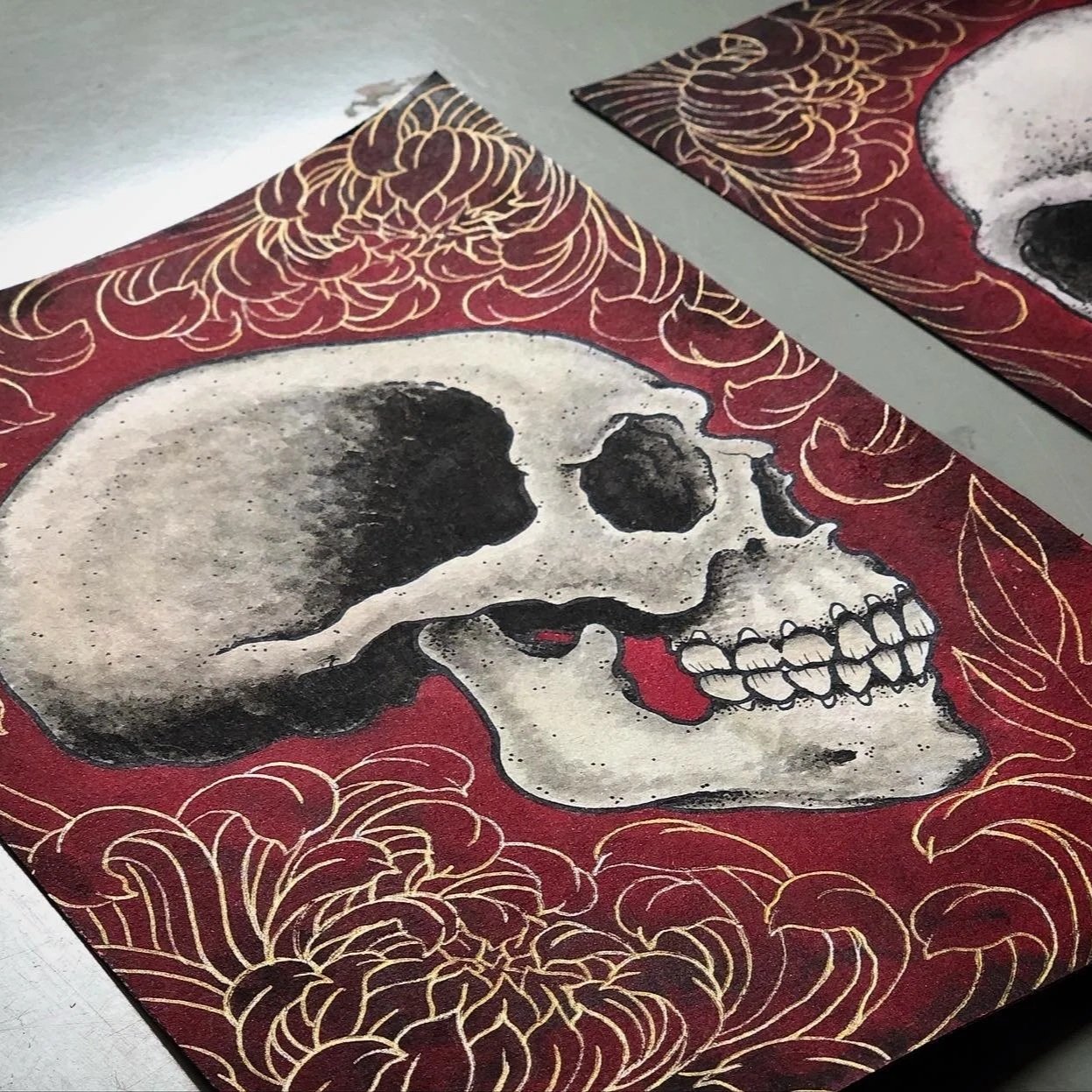 Artwork featuring a human skull illustration outlined in black and beige, set against a red background with gold line floral patterns.