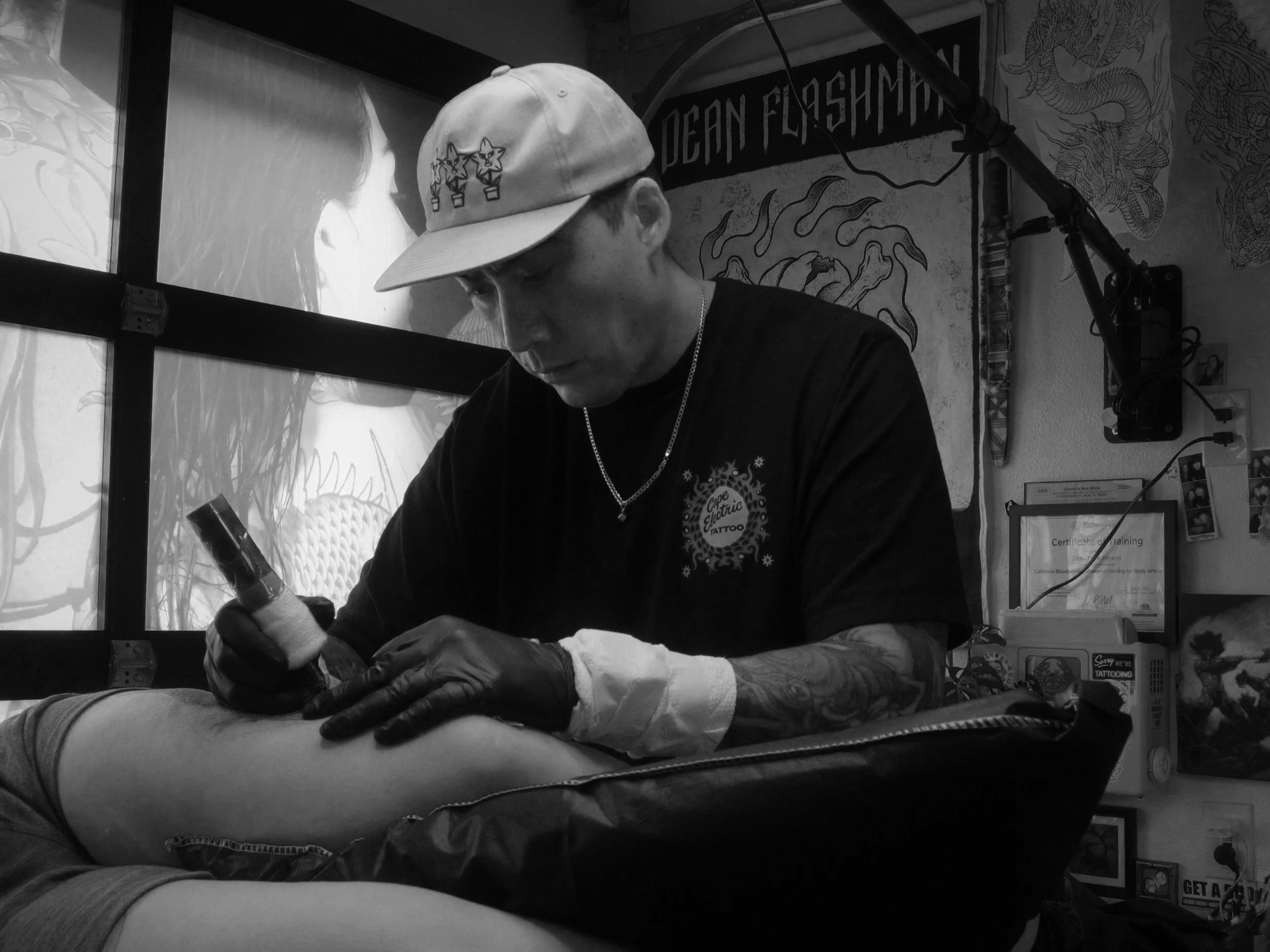 A tattoo artist working on a person's arm inside a tattoo studio. The artist is wearing a cap, gloves, and a dark T-shirt, focused on the tattooing process.