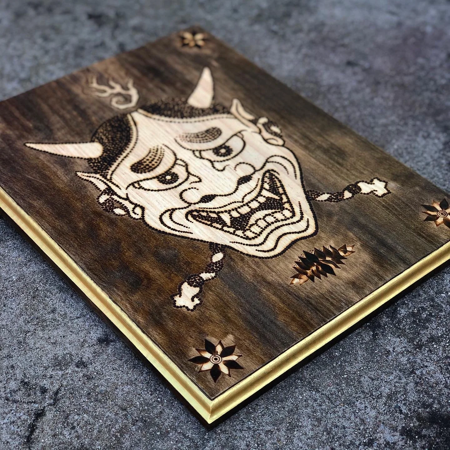 A wooden artwork depicting a cartoonish devil face with horns, wide eyes, and an open mouth. The wood has a dark stain with the devil's face carved and painted in a lighter color. Decorative leaf patterns are in each corner of the artwork.