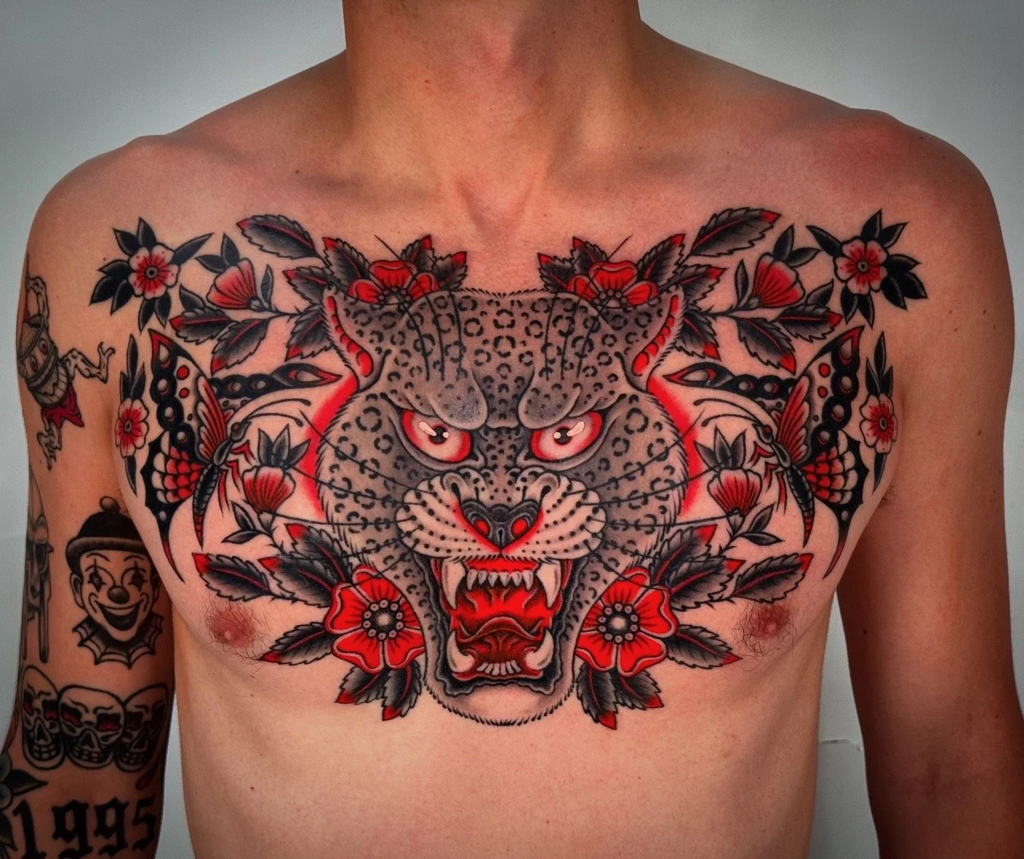 Tattoo of a fierce panther's face surrounded by flowers, leaves, and butterflies on a person's chest and arm.