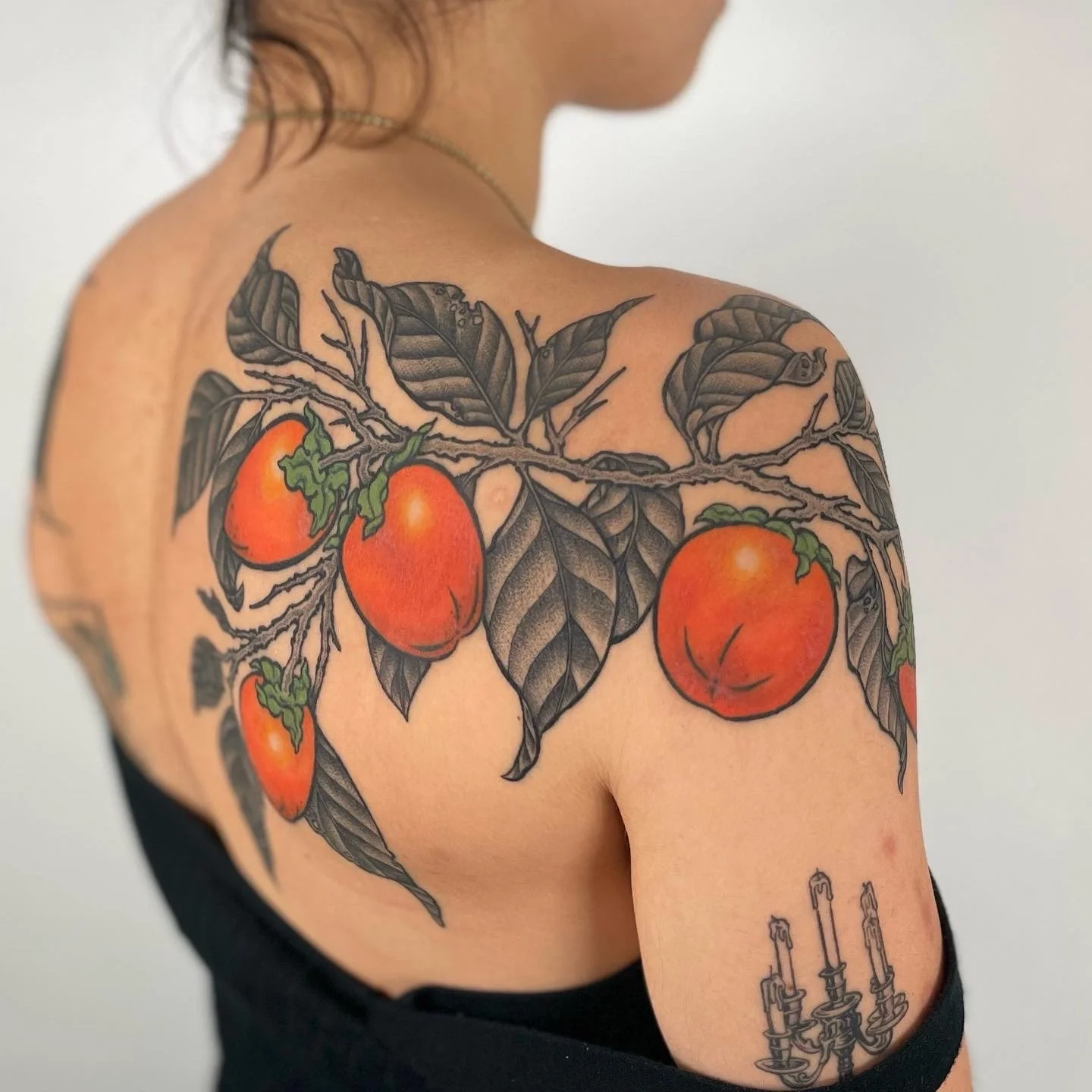 Tattoo of a branch with black leaves and three large orange tomatoes on a woman's shoulder and upper arm.
