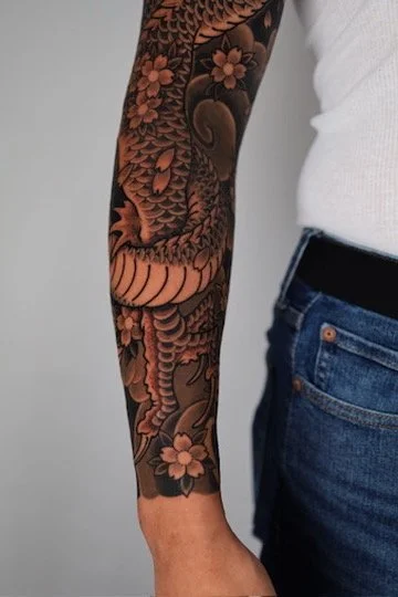 Close-up of a person's arm with a detailed black and pink floral and dragon tattoo, dressed in a white shirt and blue jeans.