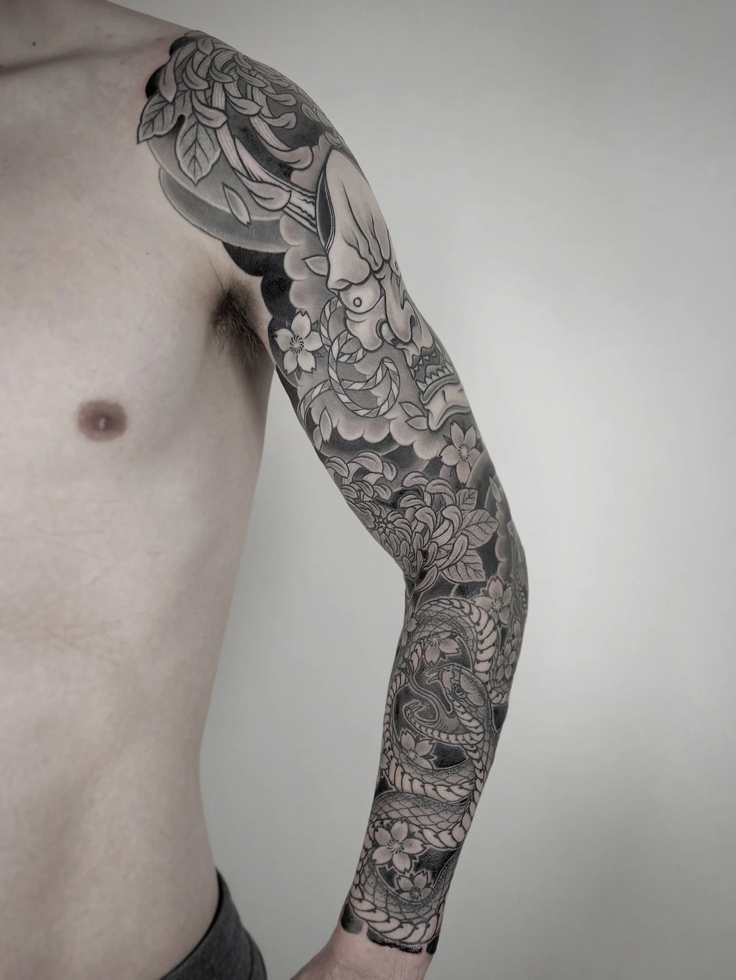 A person's left arm with a detailed black and gray Japanese-style tattoo extending from shoulder to wrist, featuring a dragon, flowers, and clouds.