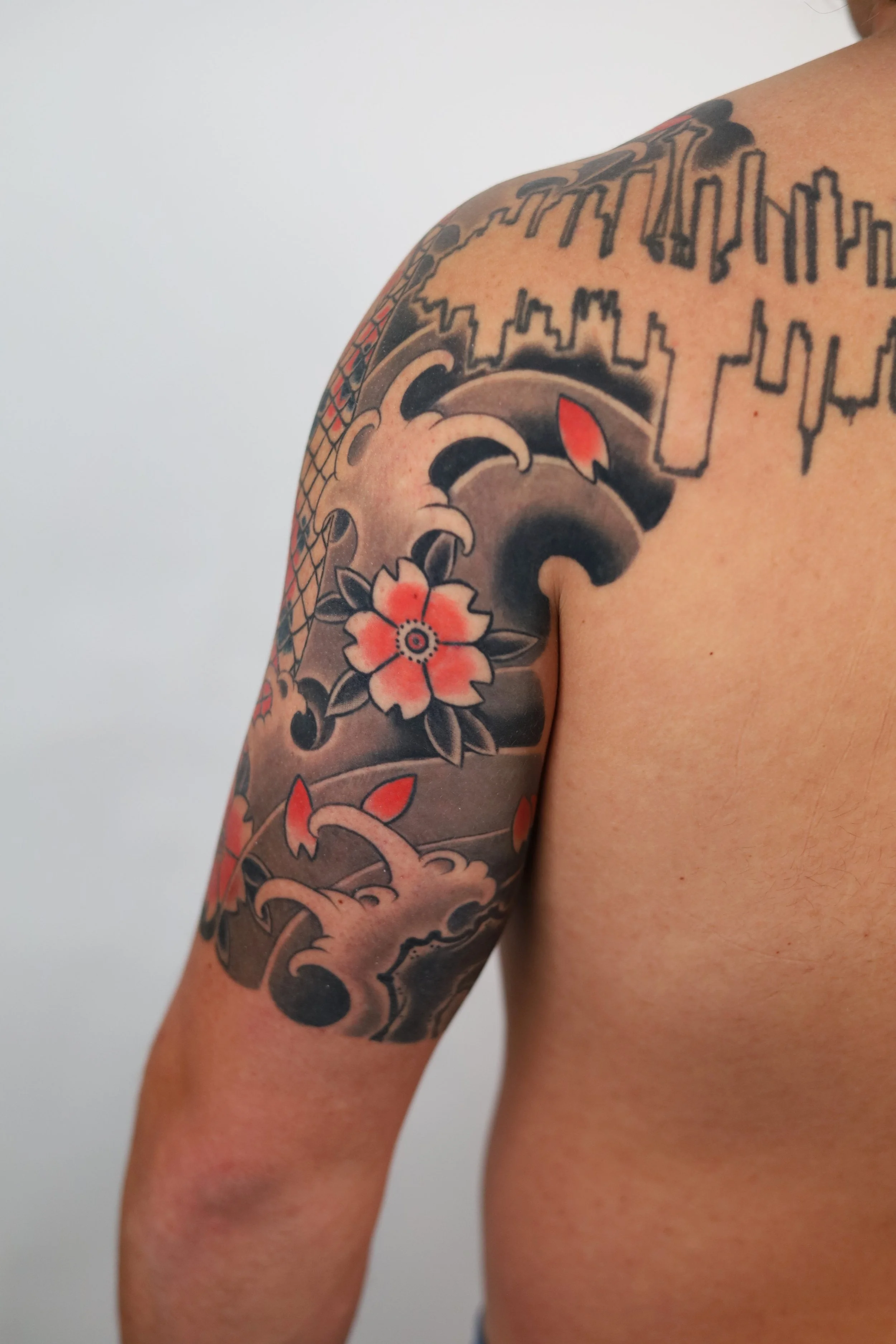 Tattoos on a person's upper arm, including a large wave, flowers, and a cityscape outline, with colors primarily in black, grey, and red.