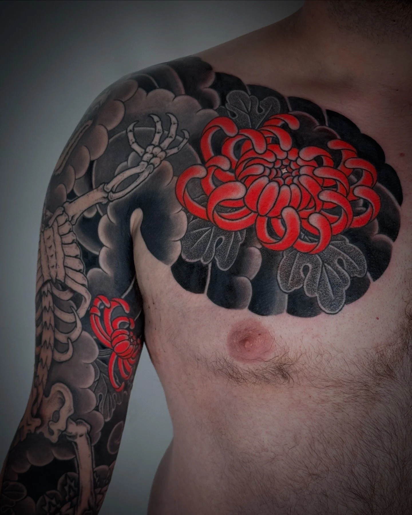 Close-up of a man's upper chest and shoulder with black and red tattoos, including a red chrysanthemum flower and a crab on the shoulder.