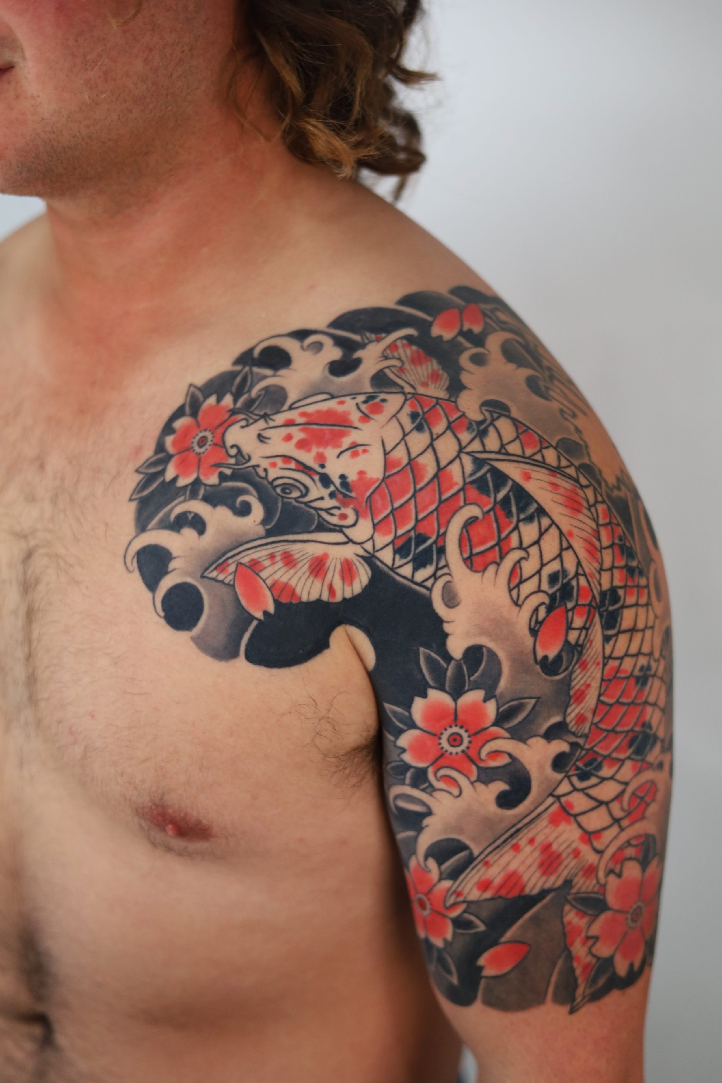 Close-up of a man's shoulder with a colorful Japanese-style tattoo featuring a koi fish, flowers, and flowing water design in black, red, and beige colors.