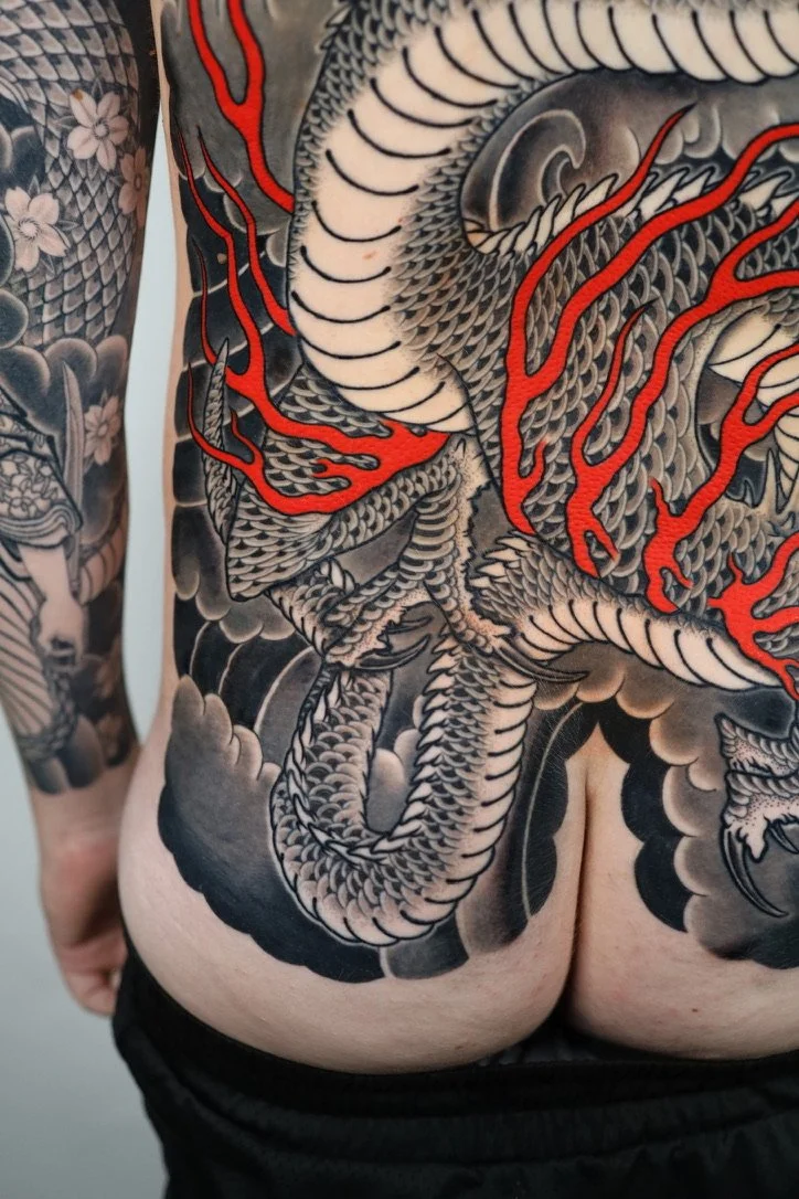 Close-up view of a tattoo featuring a detailed black and gray dragon with red accents on its flames, covering the person's back and shoulder.