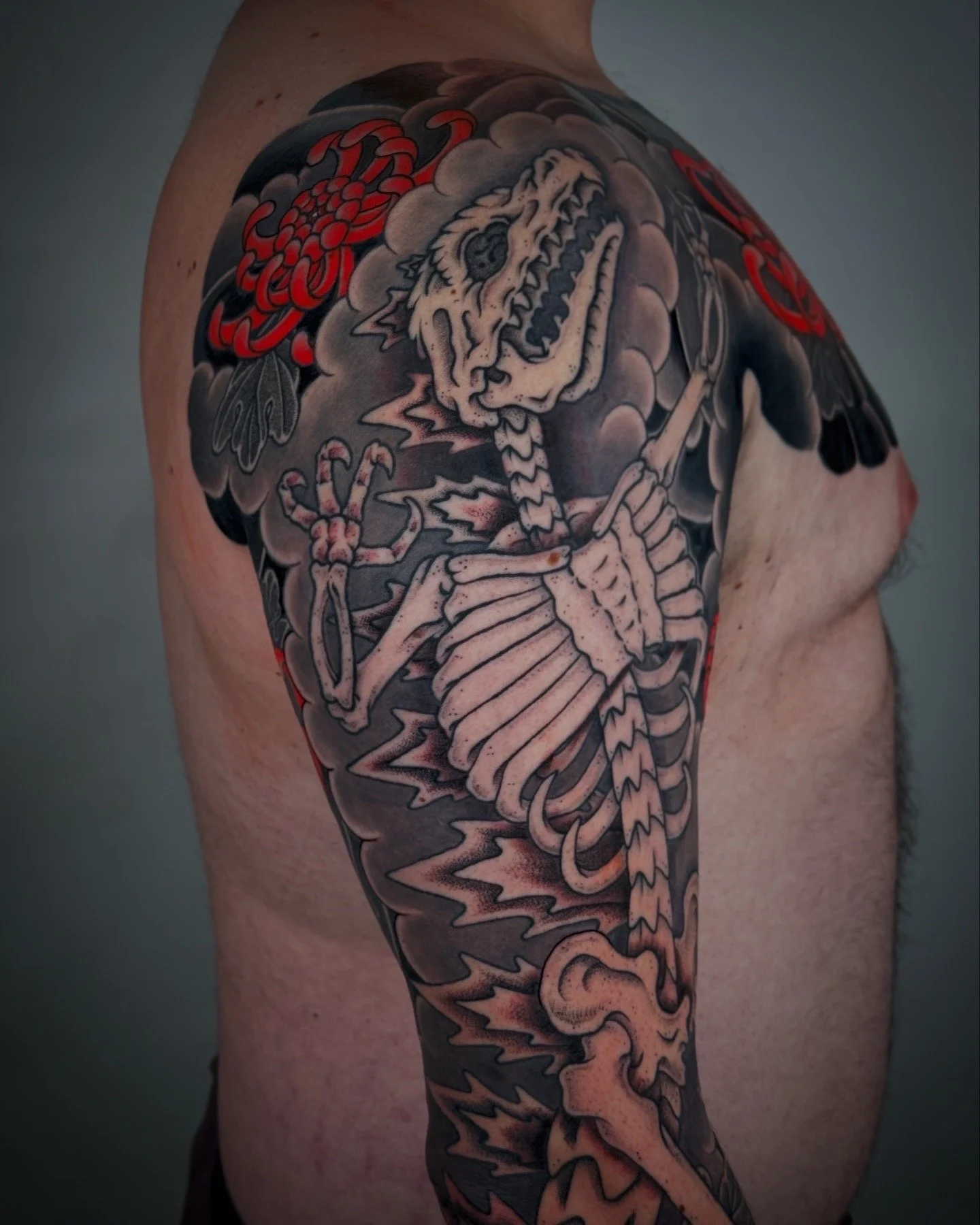 Tattoo of a skeletal dragon with a fierce skull and wings on a person's upper arm and shoulder, with dark clouds and a red flower in the background.