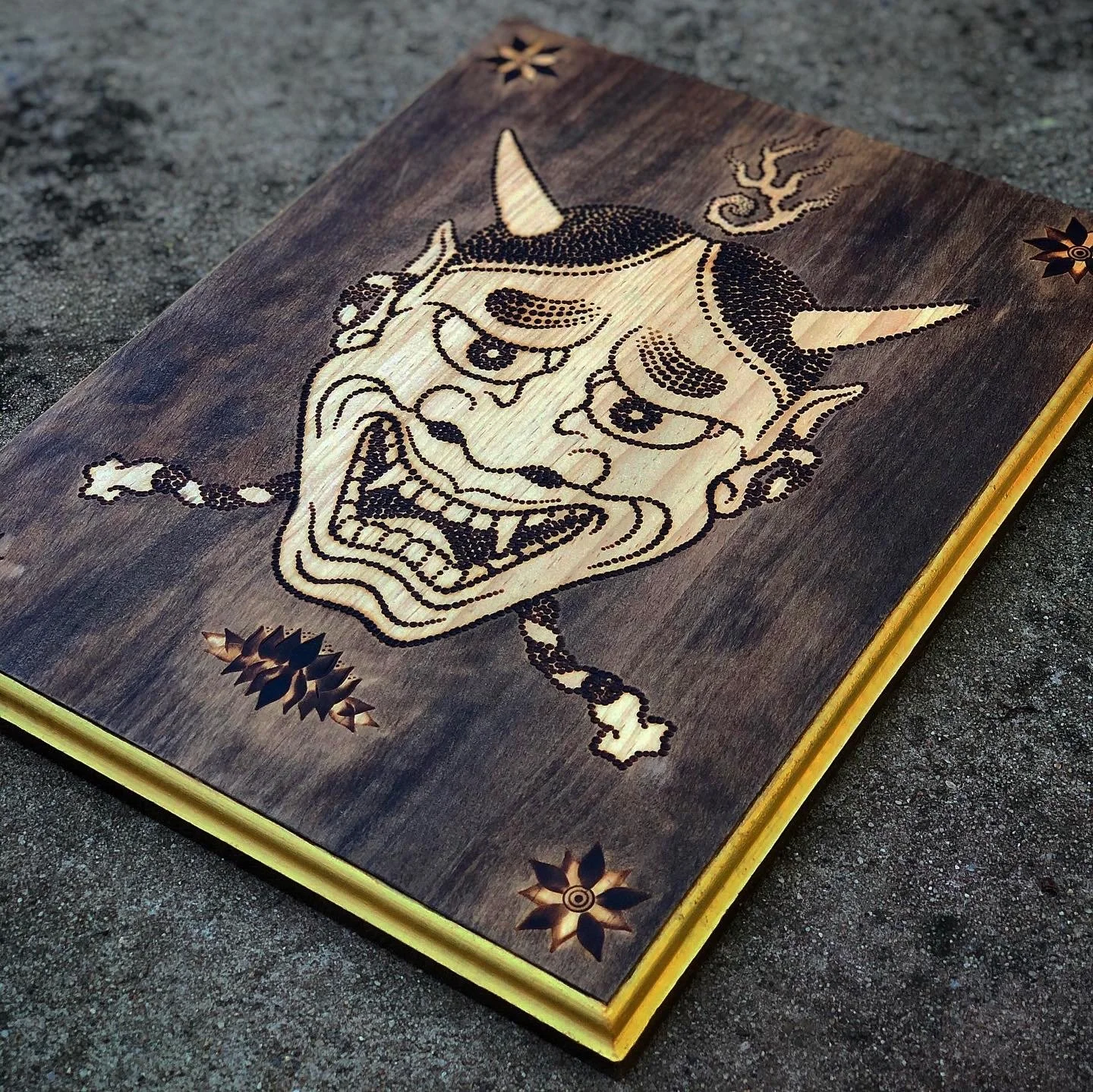 A wooden carving of a traditional Japanese demon or ogre mask, featuring detailed lines and inlays, with decorative flower inlays in each corner and a gold-colored edge.