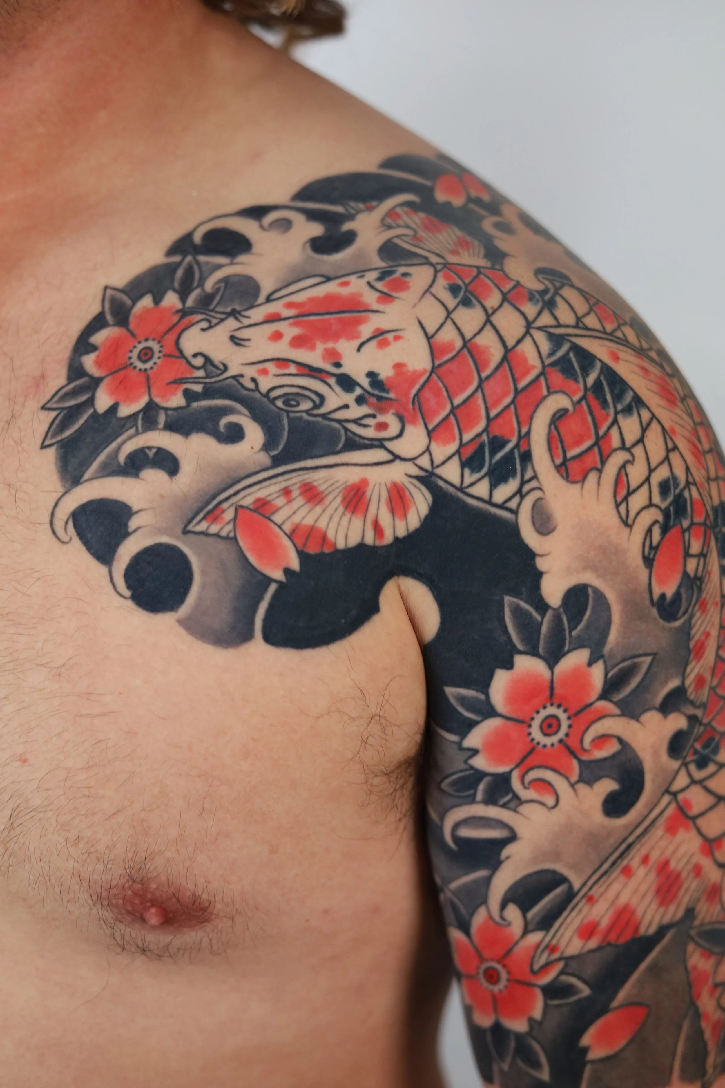 Tattoo of a koi fish with red and black accents on a person's shoulder and upper arm, surrounded by black clouds and red flowers.