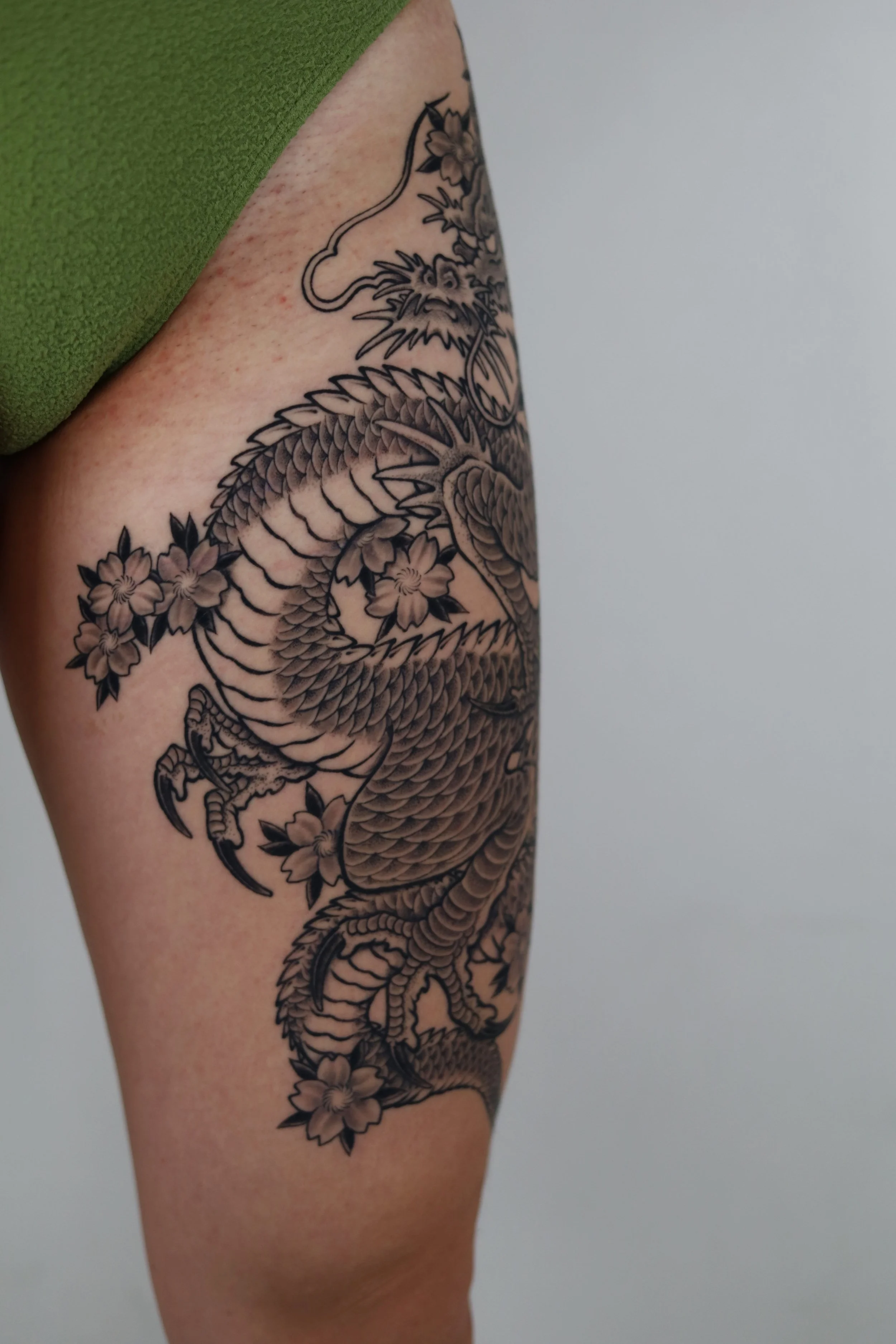 Black ink tattoo of a dragon surrounded by cherry blossoms on a person's upper thigh.