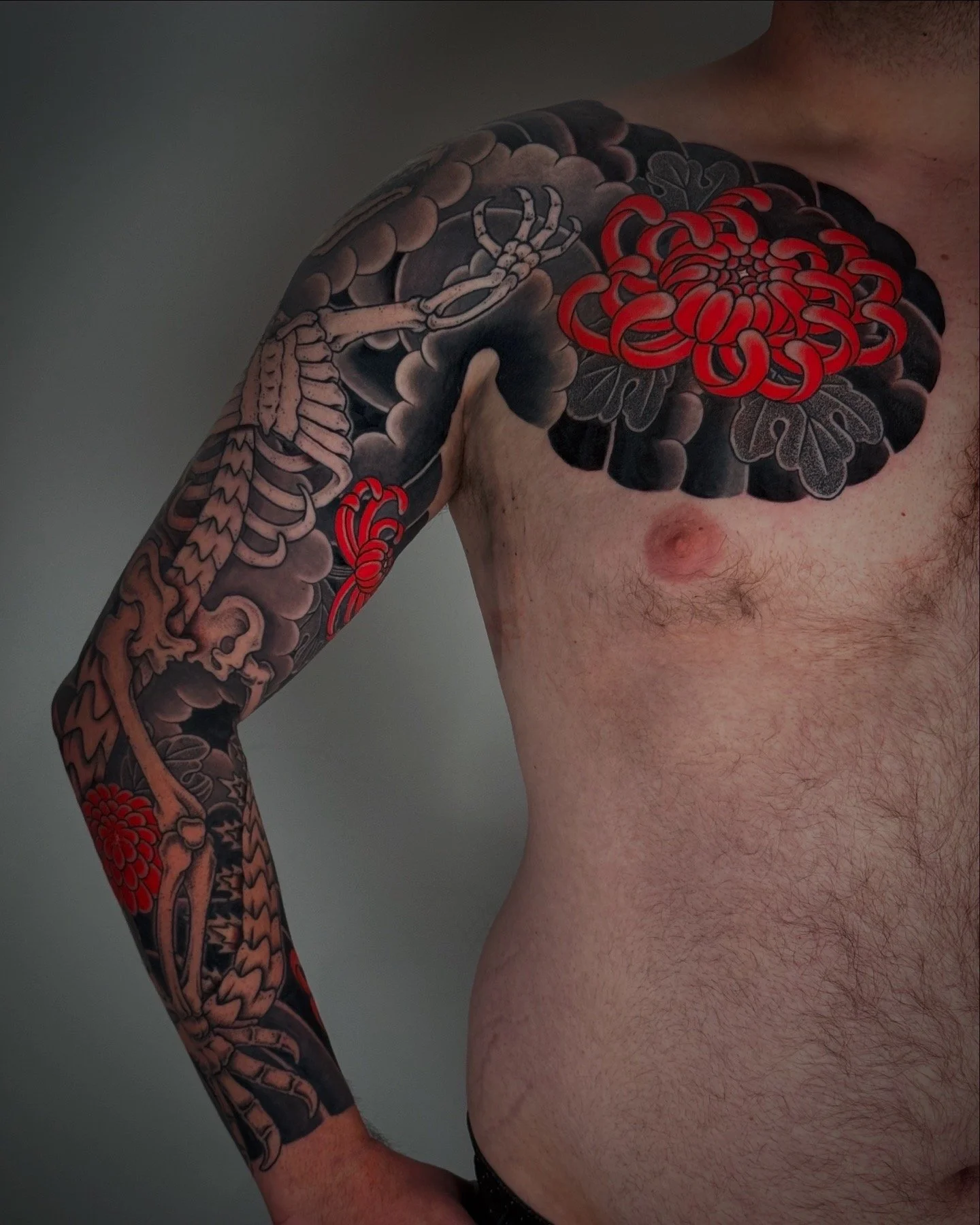 Close-up of a man's upper body showing a detailed black, gray, and red tattoo sleeve with floral and skeletal designs.
