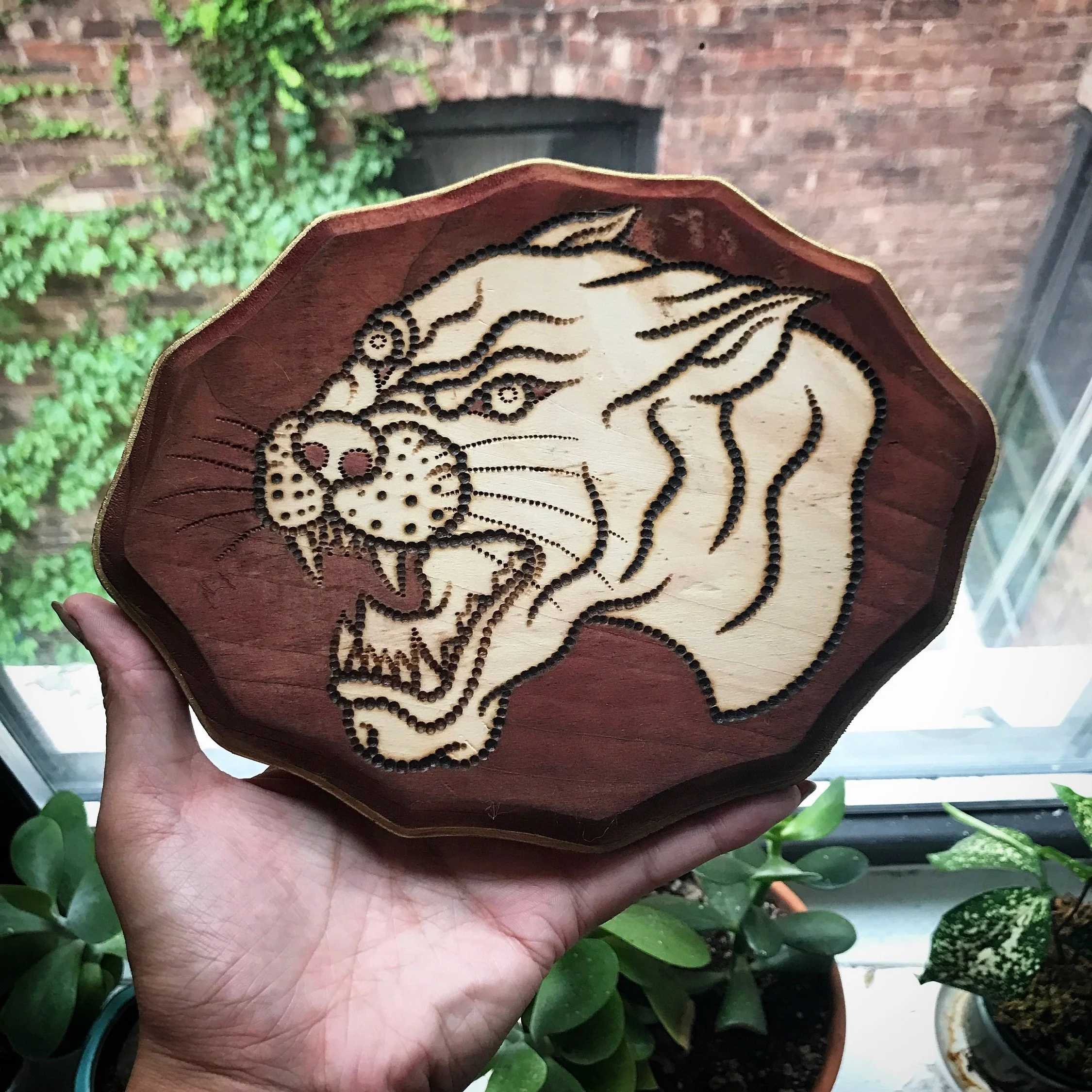 A hand holding a wooden plaque with a laser-etched image of a roaring tiger's head. The plaque has a carved, beveled edge, and the background behind it includes potted plants near a window with a brick wall outside.