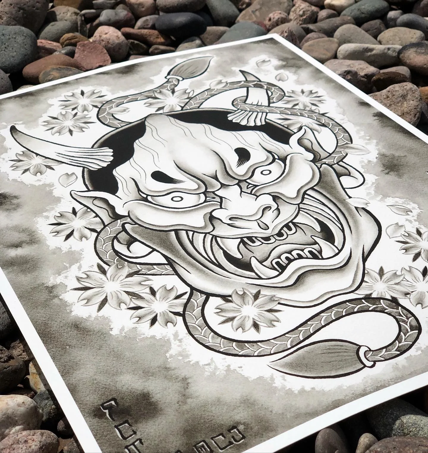 Black and white illustration of a fierce mythical creature with a demon-like face, horns, and a serpent-like body, surrounded by flowers and clouds, printed on a paper lying on rocks.