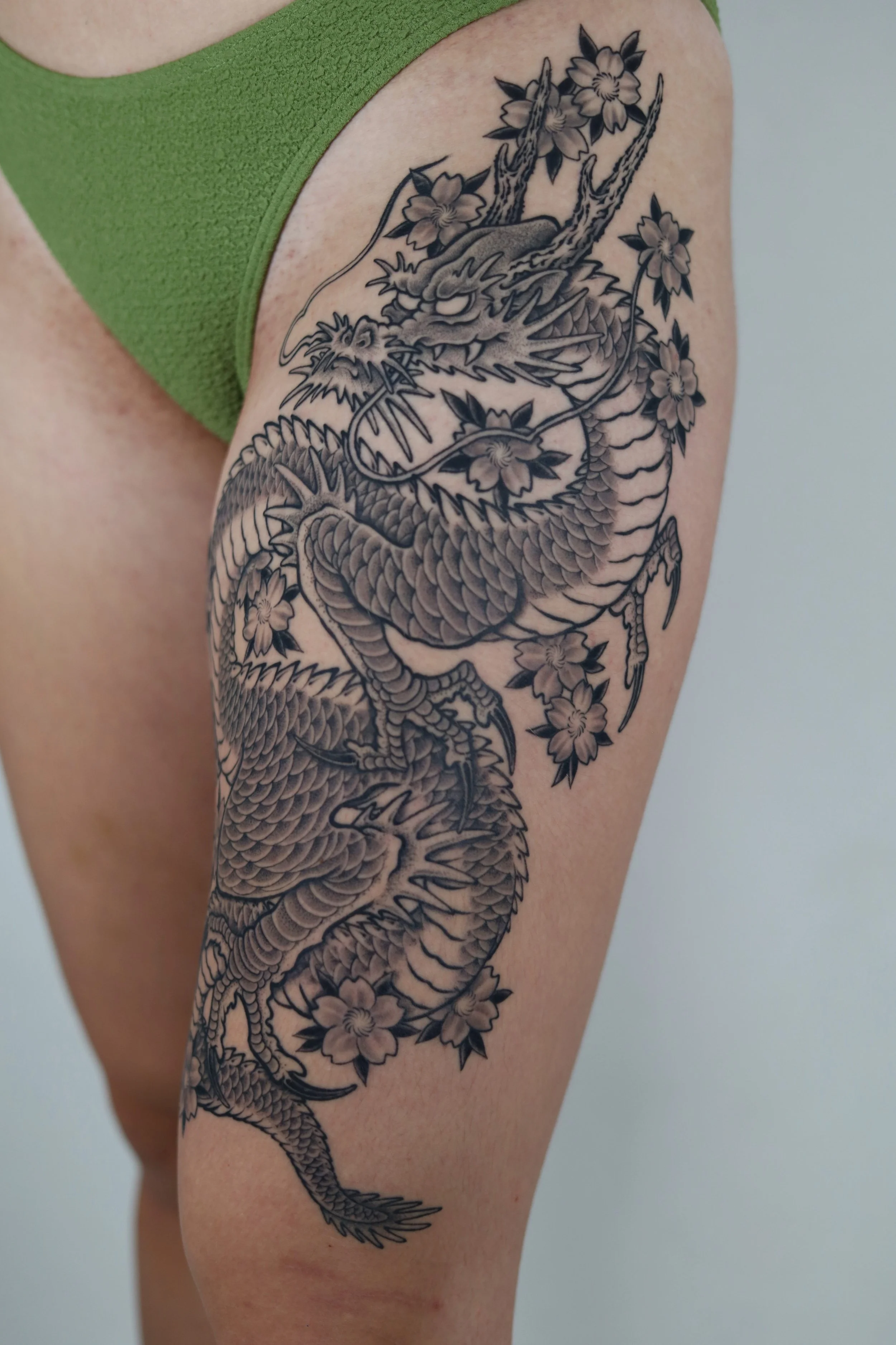 A detailed black and gray tattoo of a dragon intertwined with cherry blossoms on a person's upper thigh.