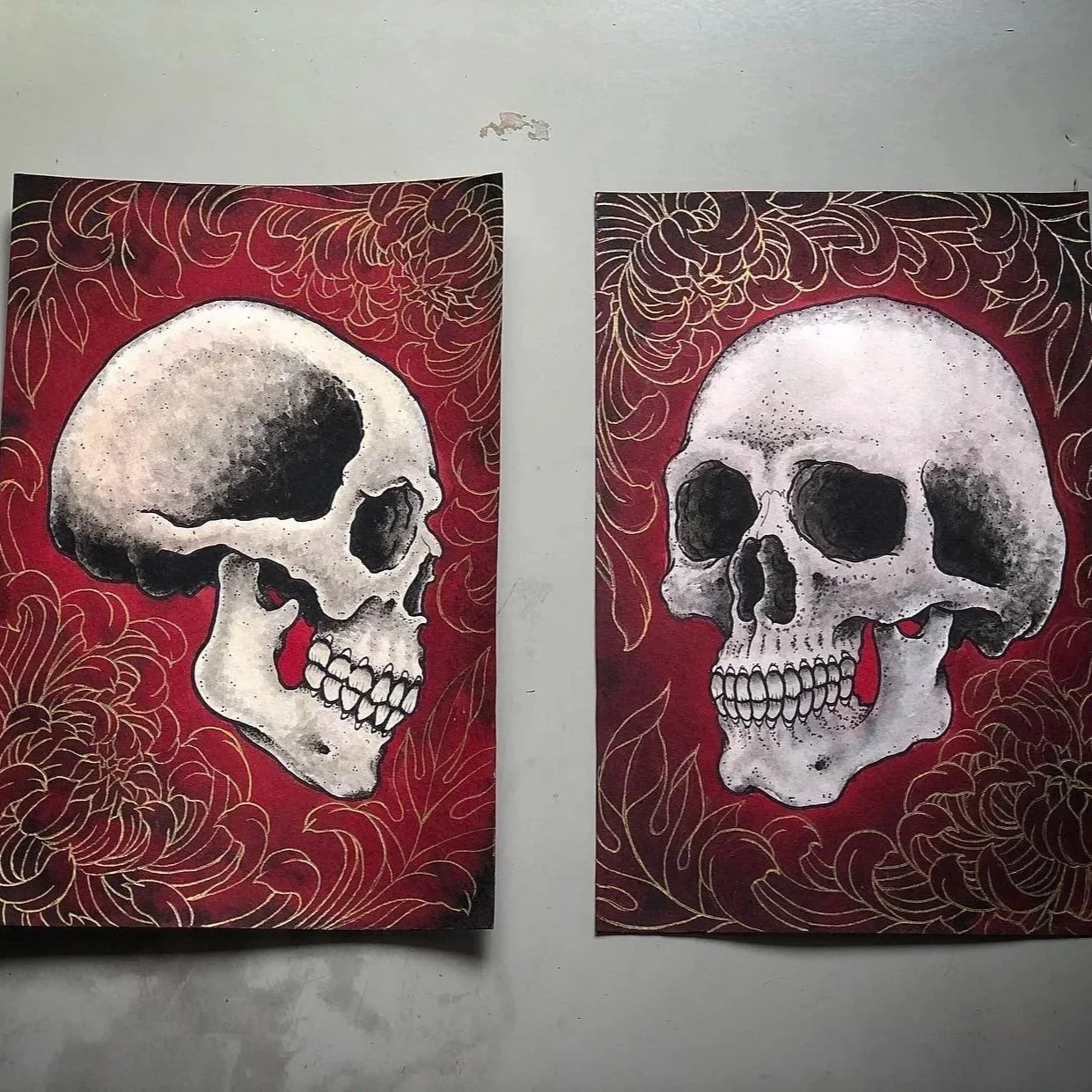 Two paintings of human skulls with floral designs on a red background, one in profile view and the other facing forward.