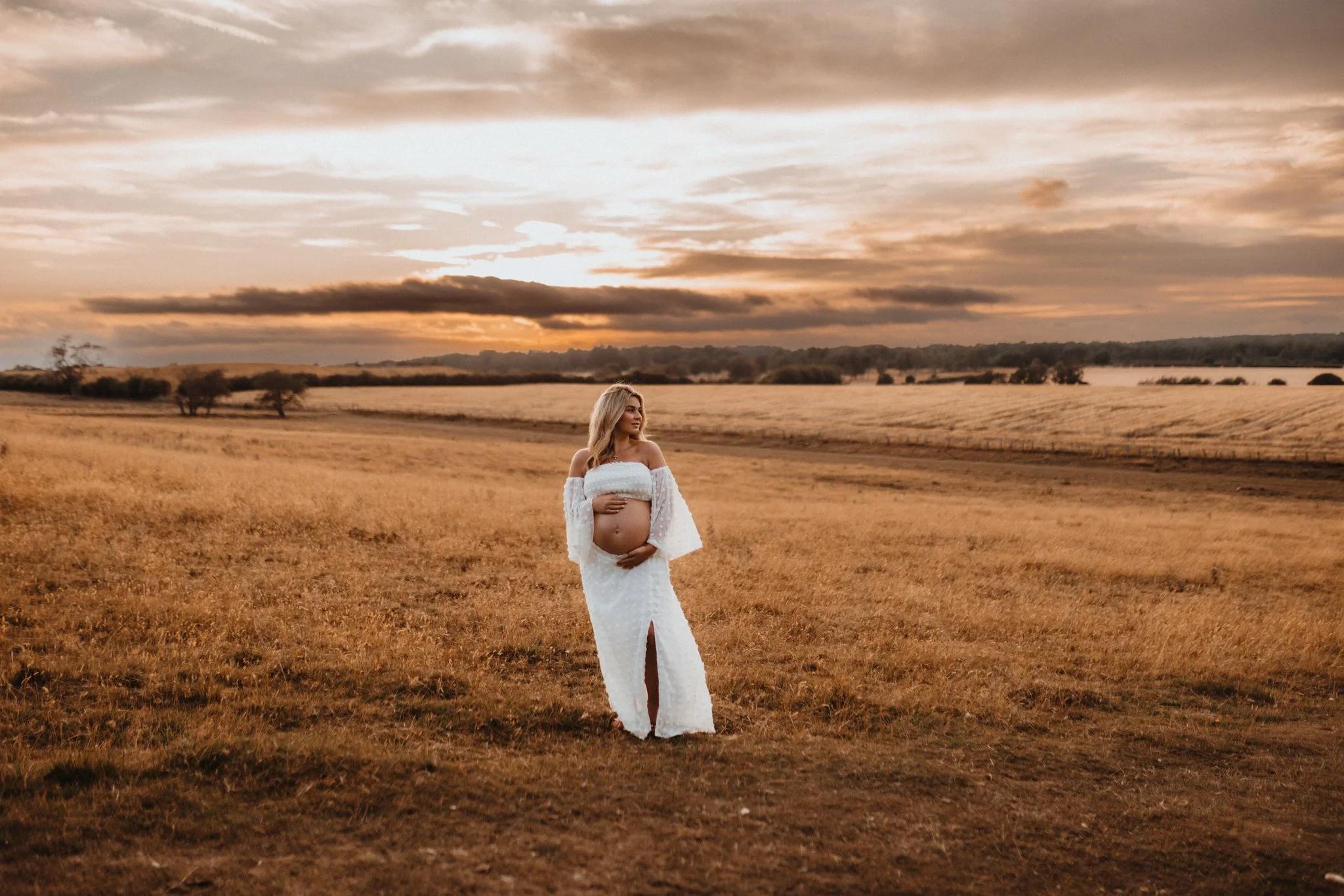 An epic maternity portrait of a pregnant woman in a white boho skirt and top standing in a field, with a sun setting behind on a summer evening in Leeds, Yorkshire