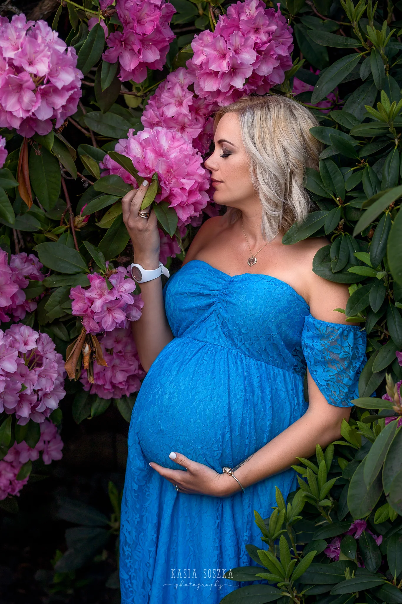 Maternity session Leeds: beautiful blond pregnant woman in a long blue dress smelling pink rhododendron flower.
