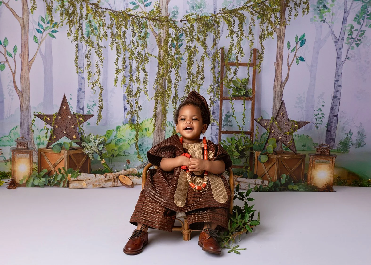 Young child dressed in traditional brown attire with orange jewelry, sitting on a small chair against a nature-themed backdrop with trees, lanterns, stars, and green foliage, smiling and holding their hands together.