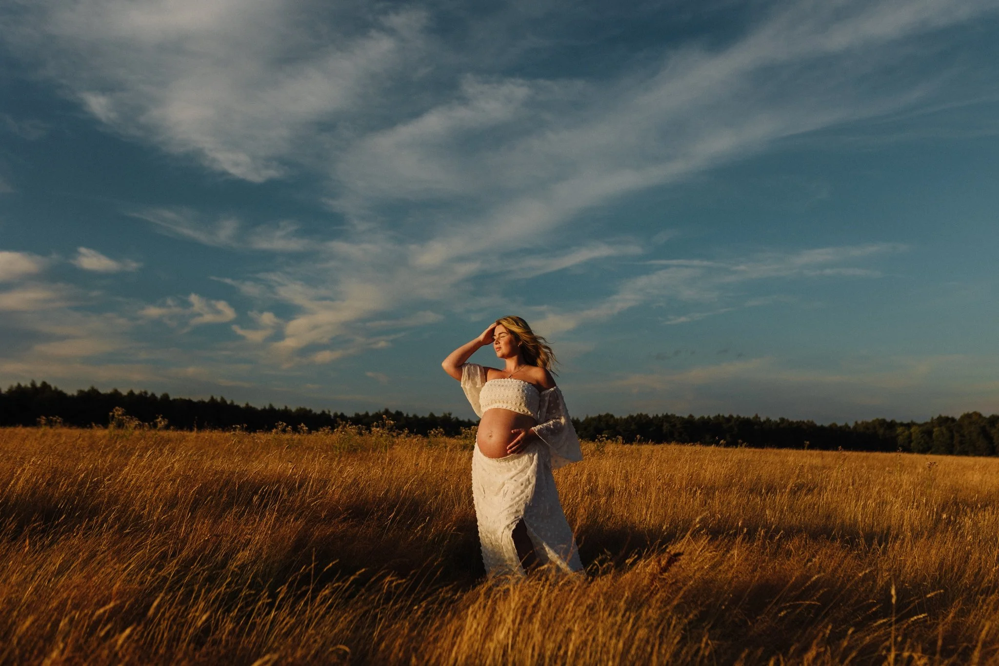 Beautiful pregnant woman in a white boho set enjoying pre-sunset golden hour glow on her face, Big blue sky, white clouds and a summer meadow. Leeds maternity photoshoot.