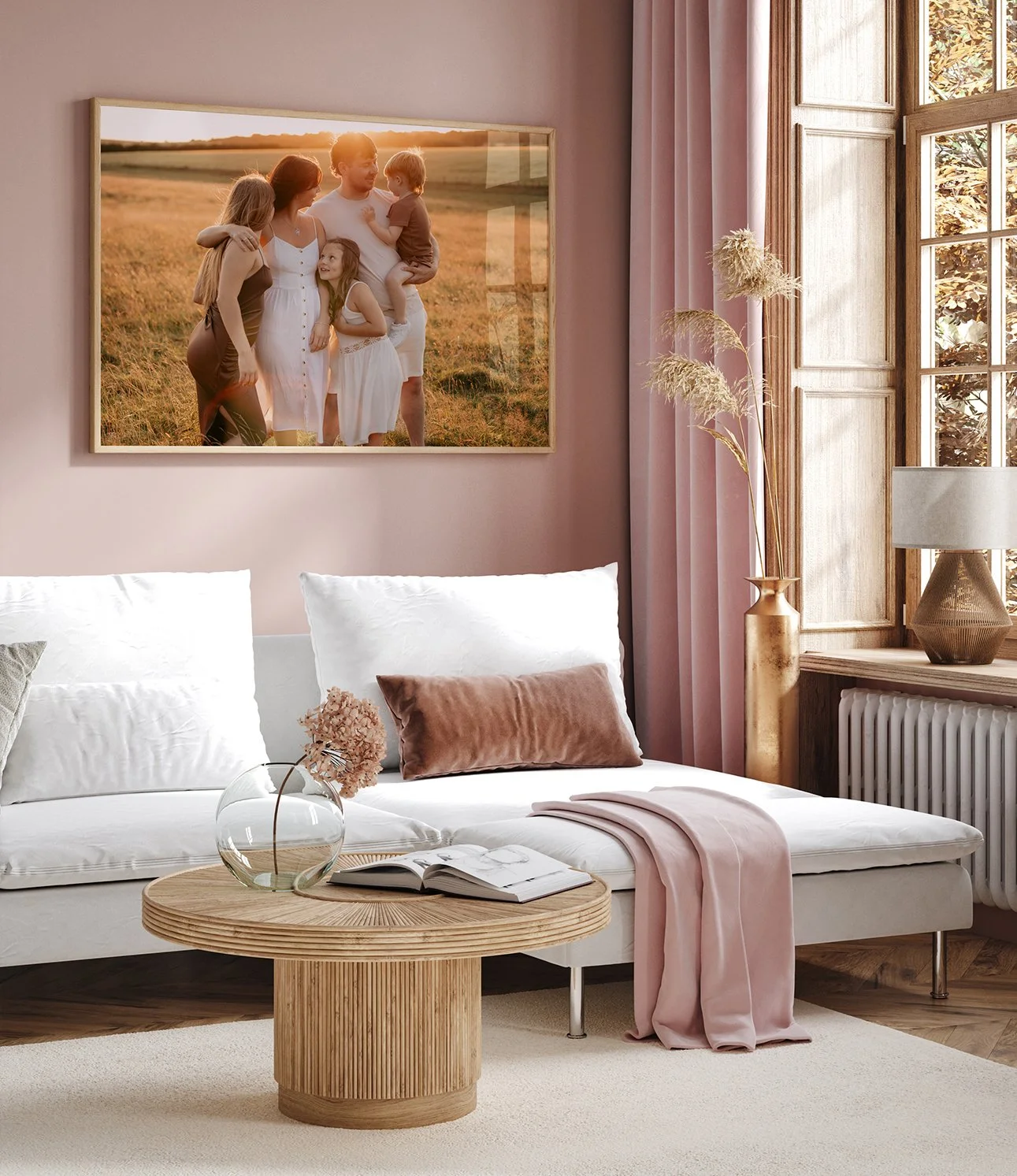 Living room with a white sofa, pink curtains, a wooden coffee table with a glass vase and an open book, framed family photograph on the wall, window with sunlight, beige and pink cushions, a pink throw blanket, and a decorative lamp and vase with dried plants.