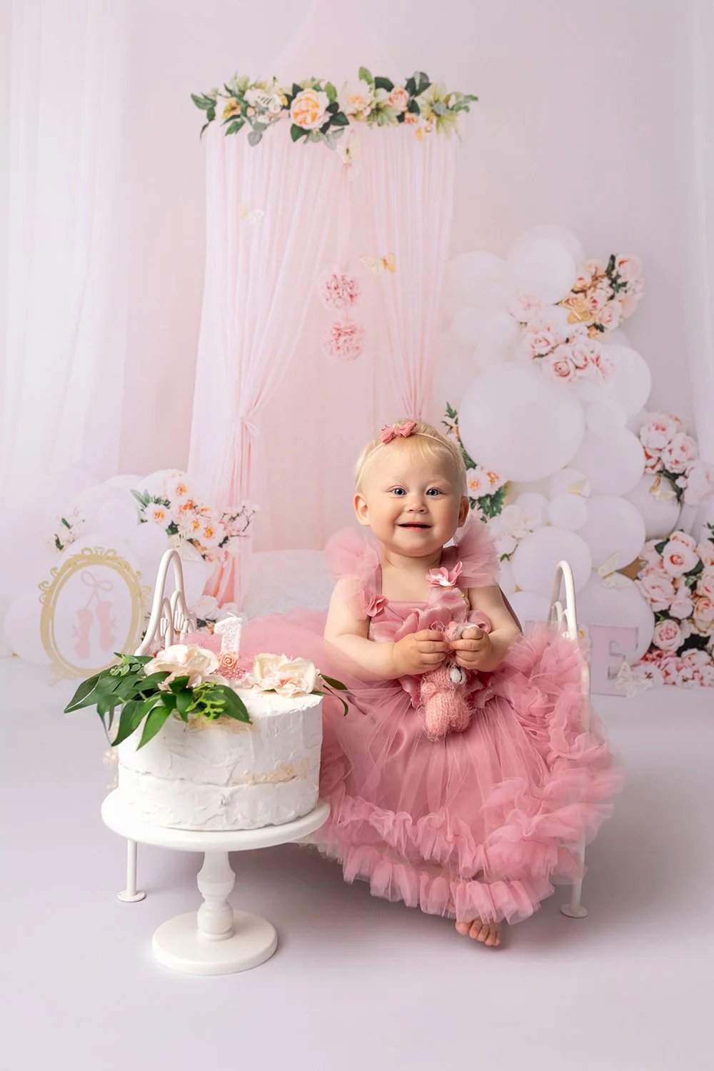 A young girl in a pink dress sitting on a small white bed with a pink tutu skirt, holding a stuffed bunny, in front of a floral background with balloons and decorative flowers, with a white cake with pink flowers and greenery in front of her.