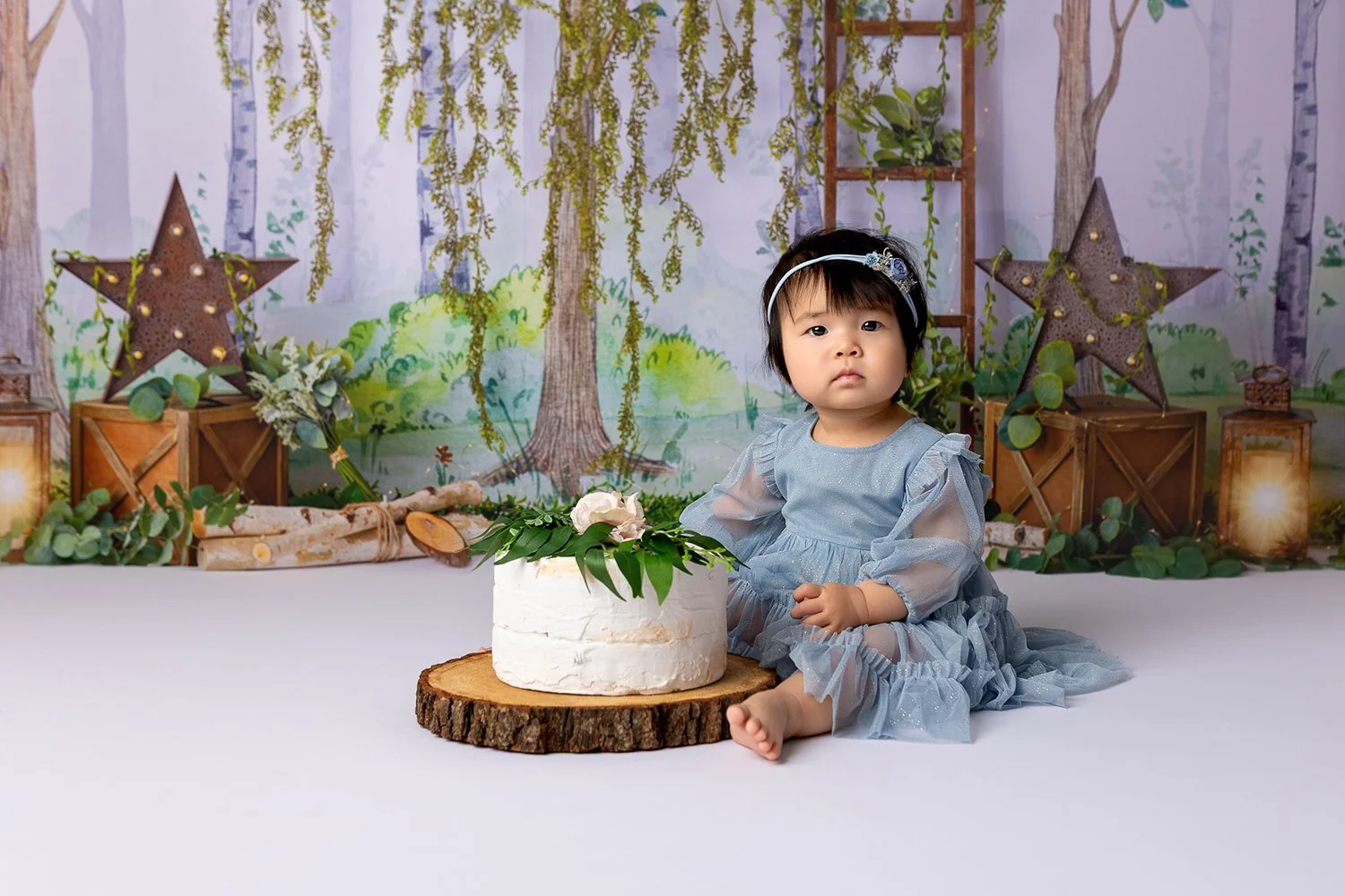 A young girl in a gray dress is sitting on the floor in front of a decorated backdrop with woodland and nature themes, next to a white cake decorated with flowers and green leaves, on a wooden slab.