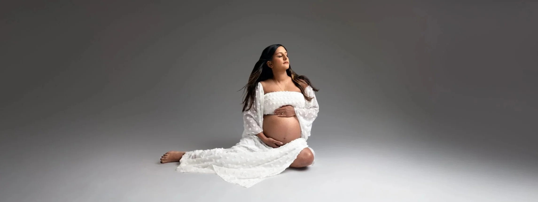 A pregnant woman in a boho crop top and a maxi skirt sitting on a floor with eyes closed during her maternity session in Leeds
