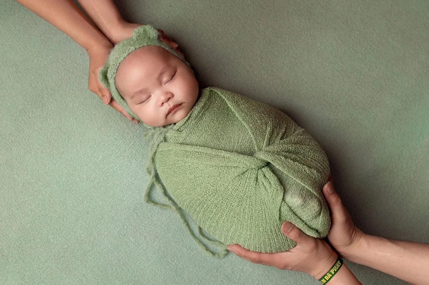A sleeping baby wrapped in a green blanket with a matching hat, lying on a green surface, with an adult's hands gently supporting the baby's head and feet.
