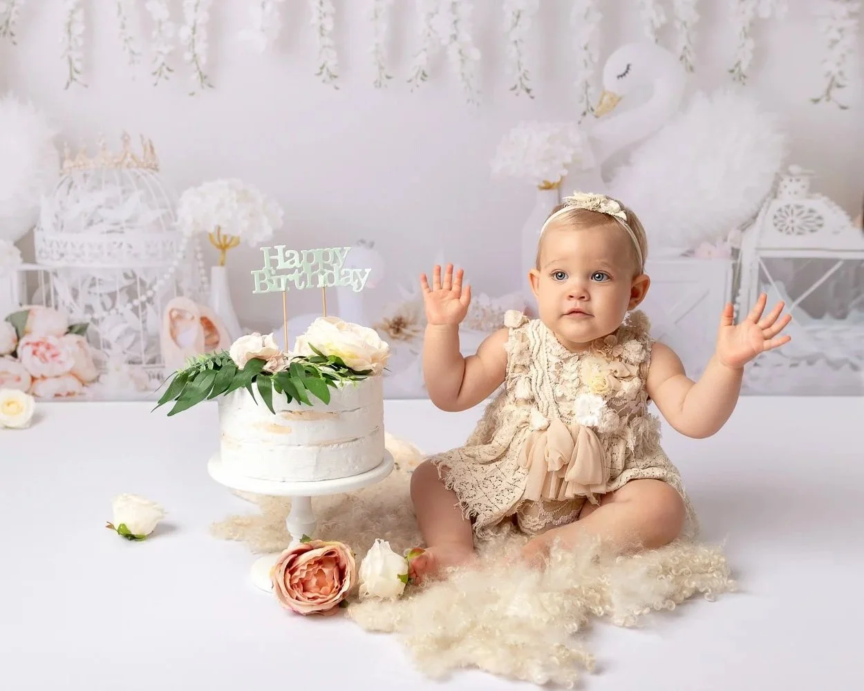 A baby girl sitting on a soft blanket in a decorated room for her birthday. She is wearing a cream lace dress with a bow, with her hands raised. In front of her is a white birthday cake with green leaves, flowers, and a 'Happy Birthday' topper. Flowers are scattered around her and the background features elegant white decor and a swan figure.