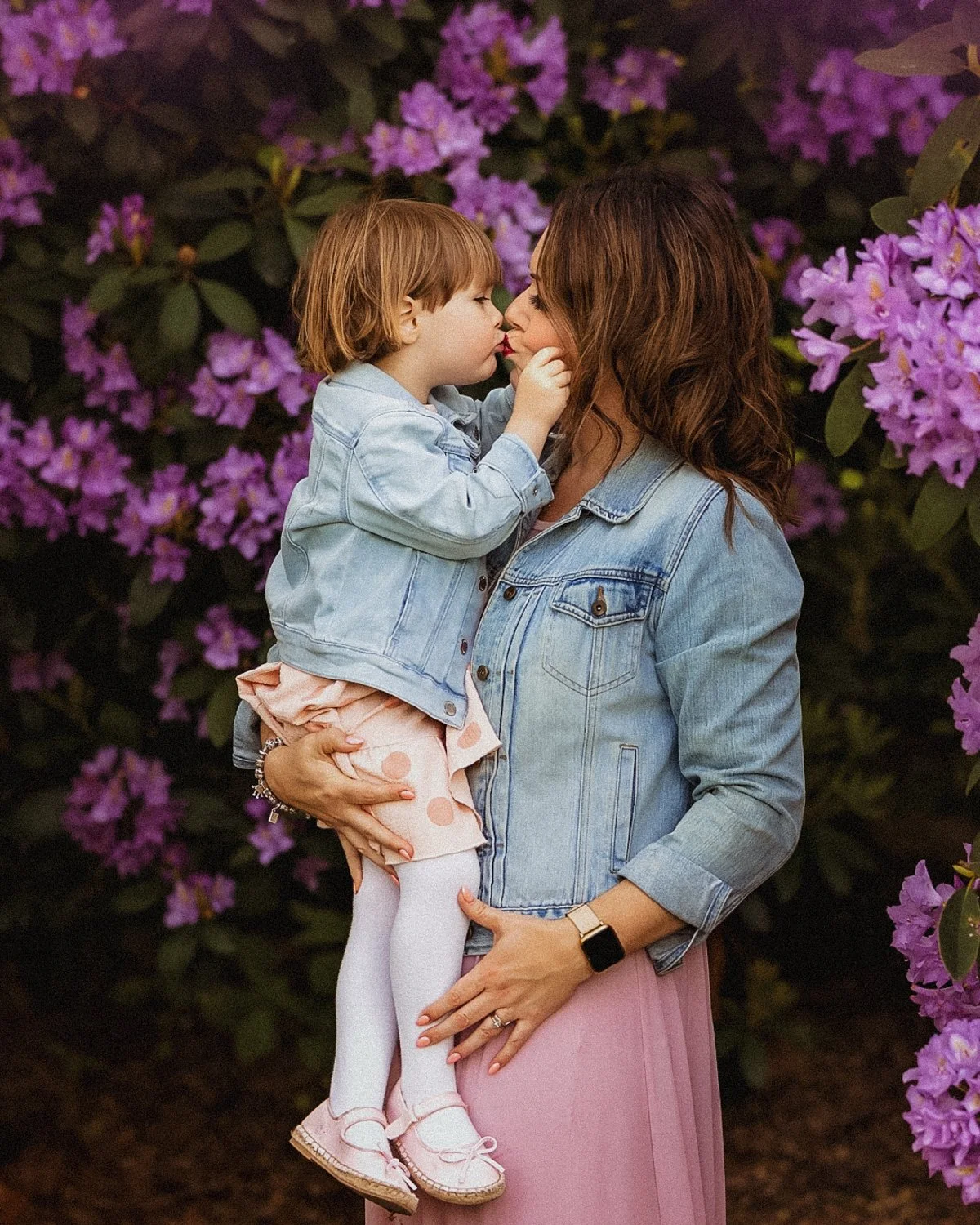 Mother kissing her little daughter. Both dressed in blue denim jackets, surrounded by blossoming rhododendrons. 