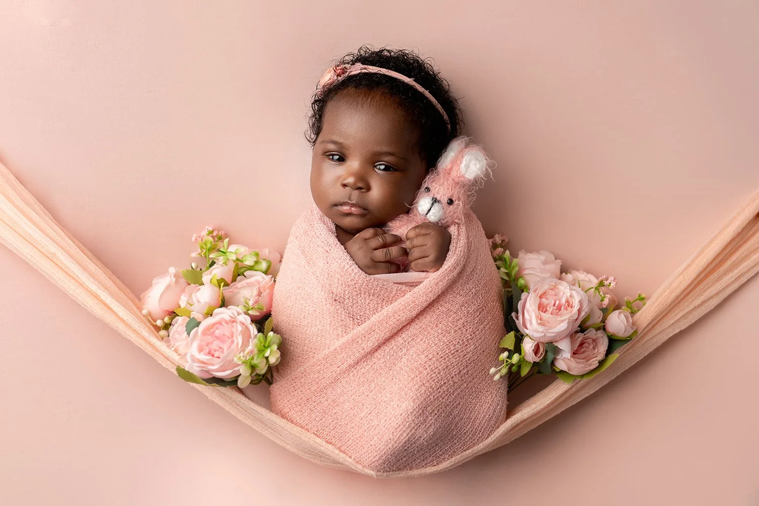 A baby girl wrapped in a pink blanket, holding a pink stuffed bunny, surrounded by pink flowers on a soft pink background.