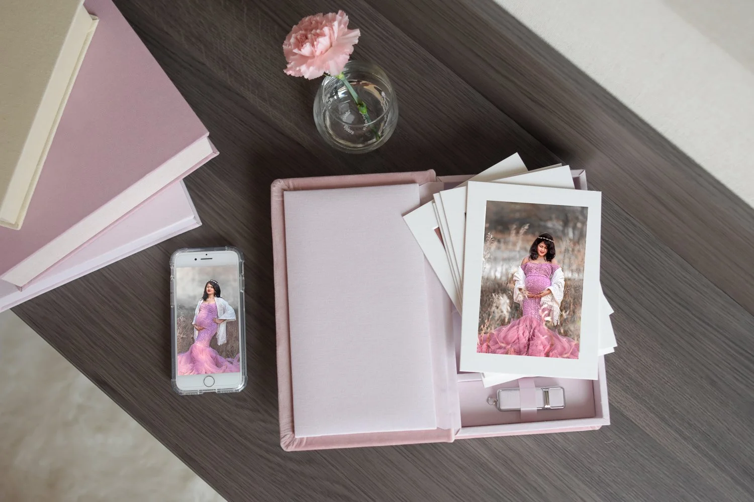 A pink box filled with stacked photographs of a woman in a pink dress and a white jacket, a single print outside the box, a smartphone showing the same woman, a pink flower in a small vase, and a couple of pink books on a wooden table.