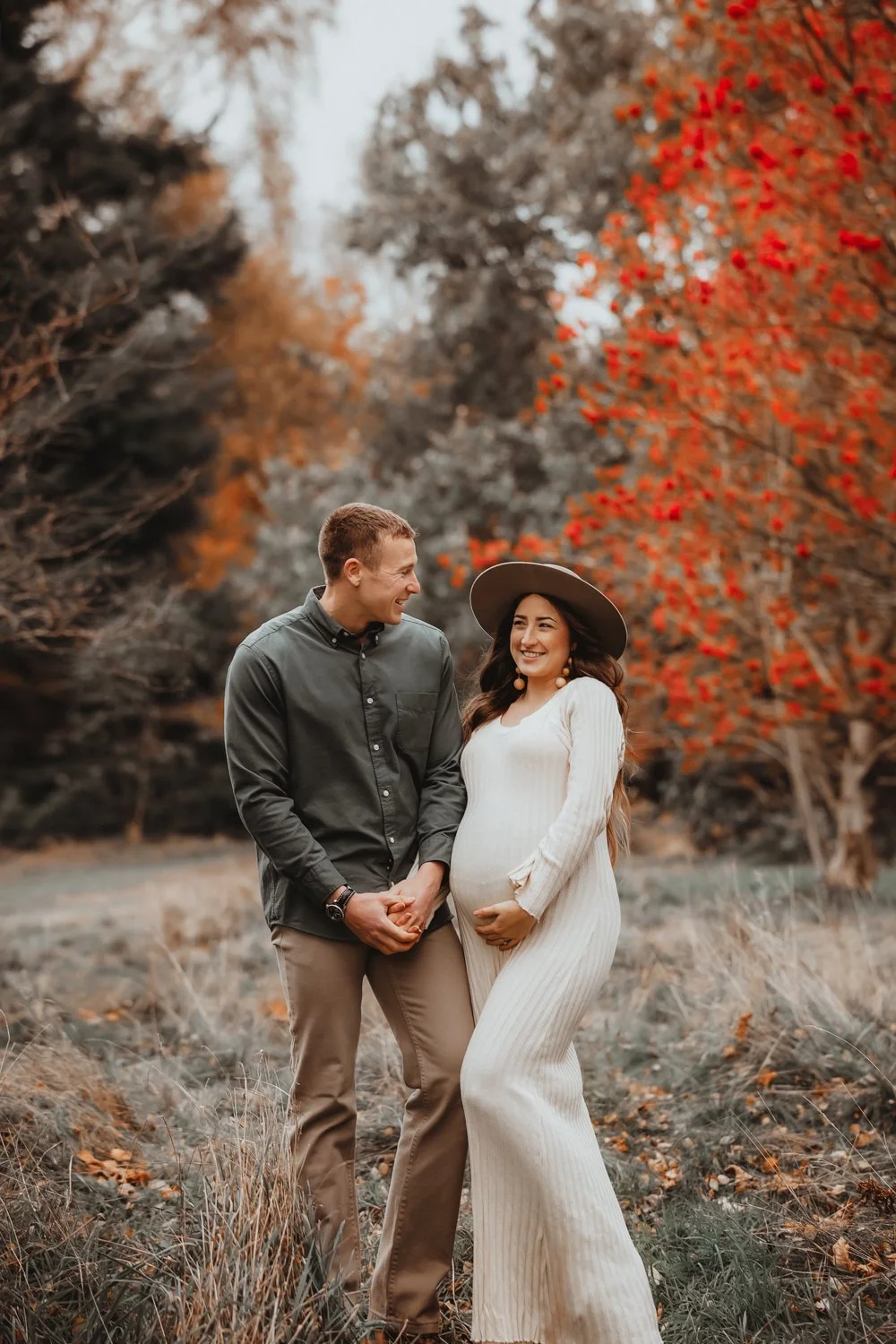 Pregnant woman in a long sweater dress and a fedora hat smiling at her husband during their maternity photoshoot on an autumn day in Leeds.