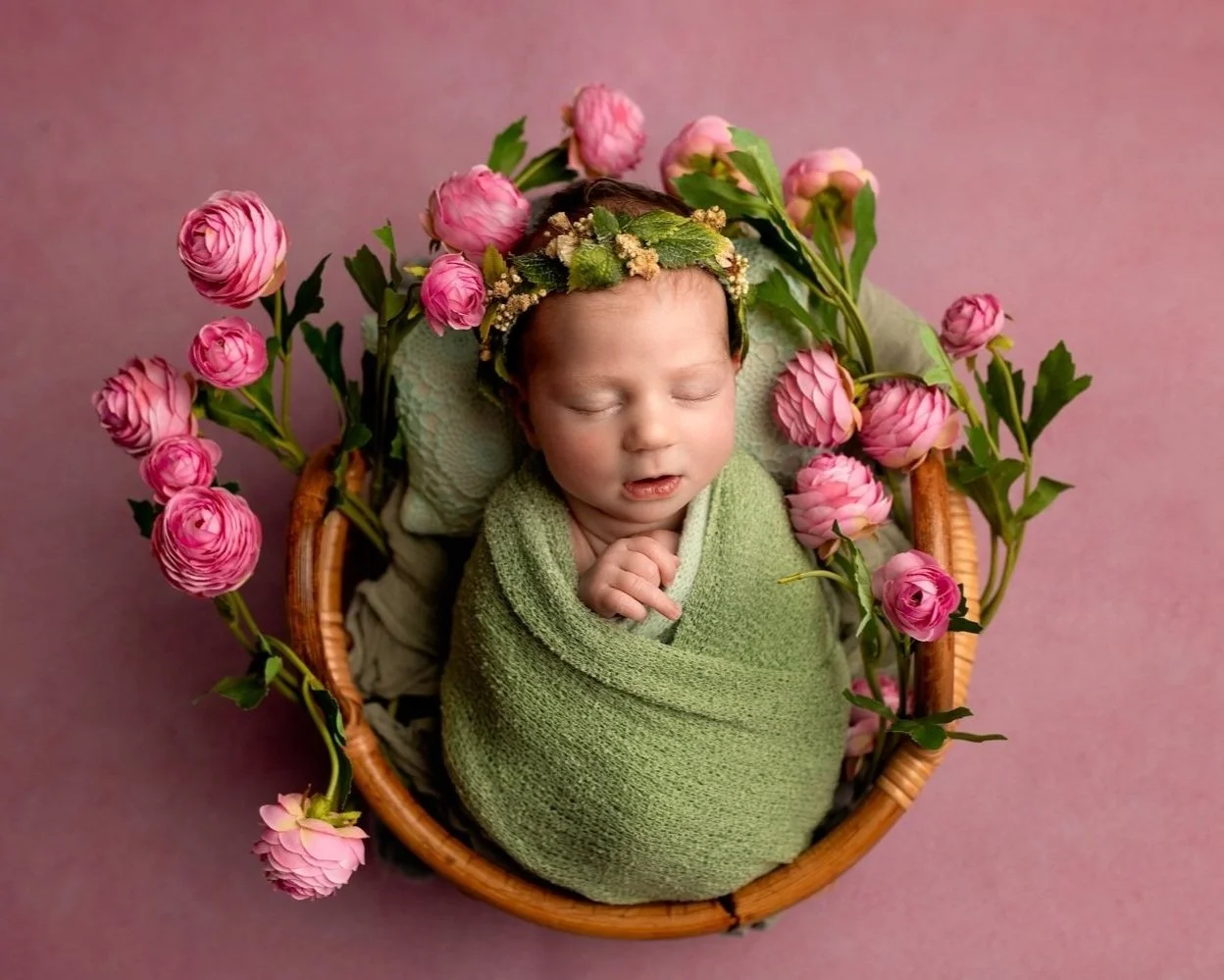 Newborn baby girl wrapped in a green fabric and posed in a basket surrounded by pink flowers