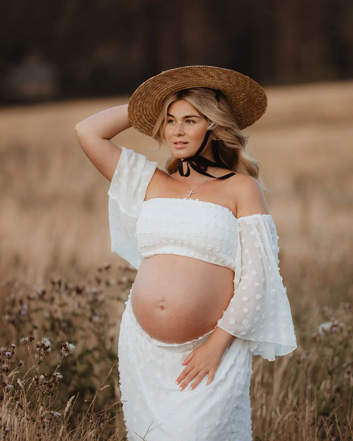 Pregnant woman in a white top, a maxi skirt and a straw hat standing in a summer meadow during her maternity shoot in Leeds