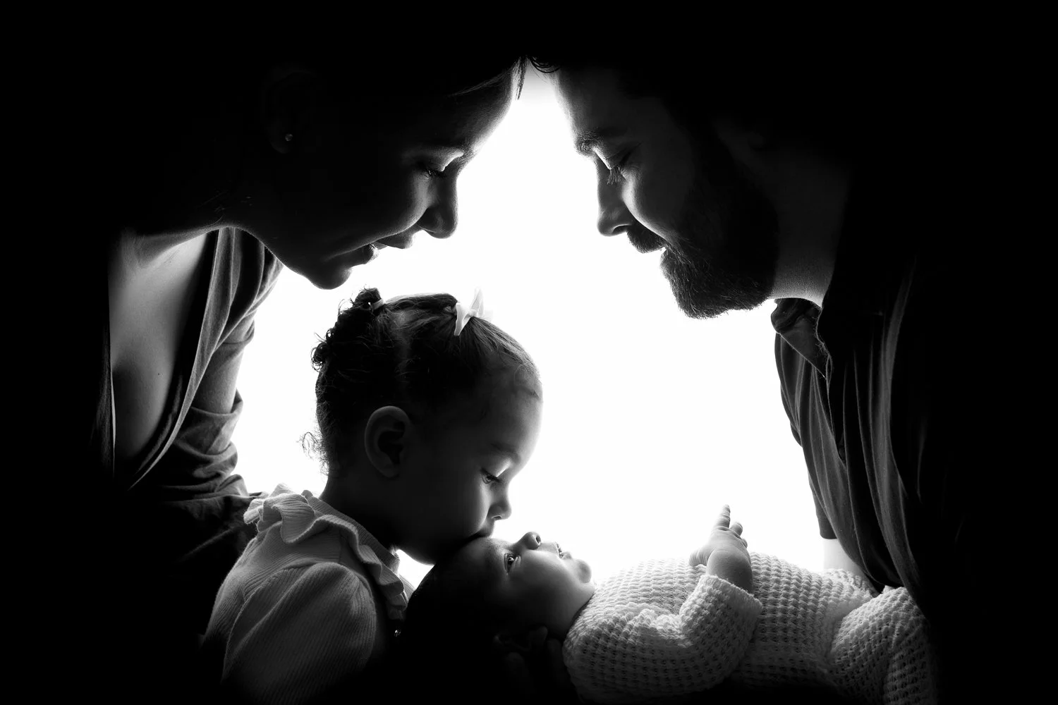 A backlit black & white portrait of mum, dad, and their little daughter kissing her newborn baby boy