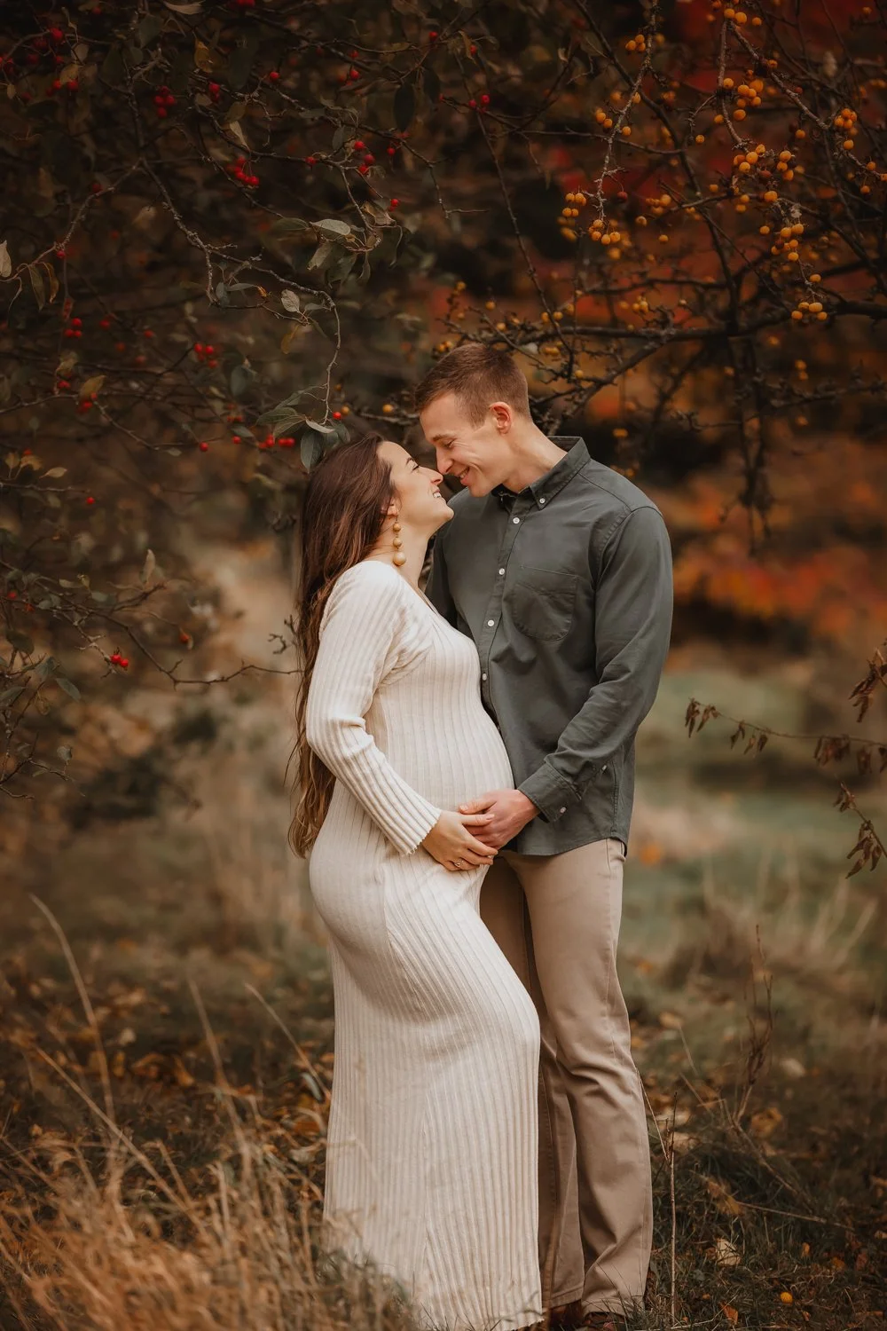 Pregnant woman in a cream sweater dress sharing a sweet moment with her husband during their maternity photoshoot in Leeds. They are looking at each smiling, surrounded by autumn colours of Golden Acre Park