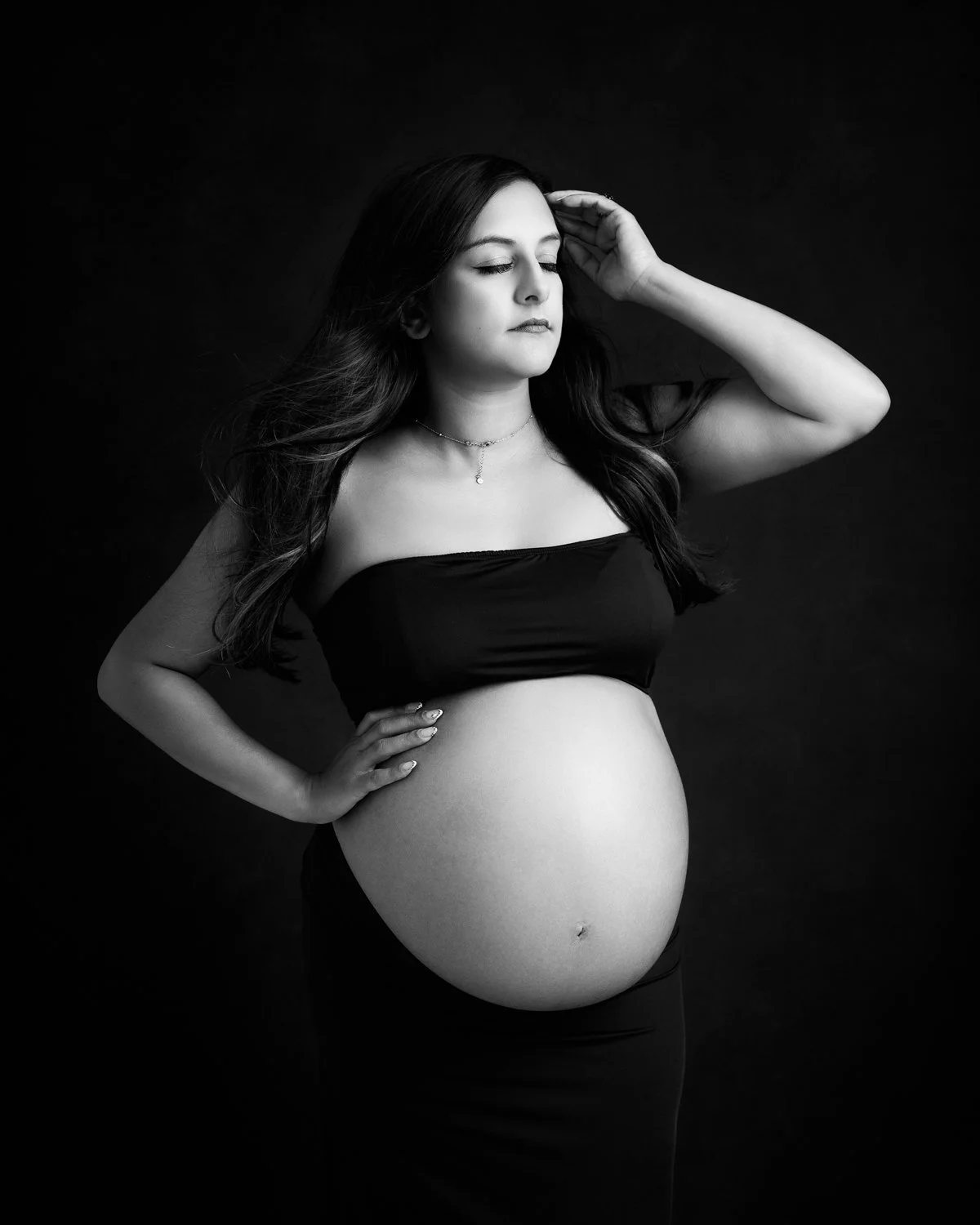 A black& white portrait of a pregnant woman with long dark hair wearing a cropped top and a skirt