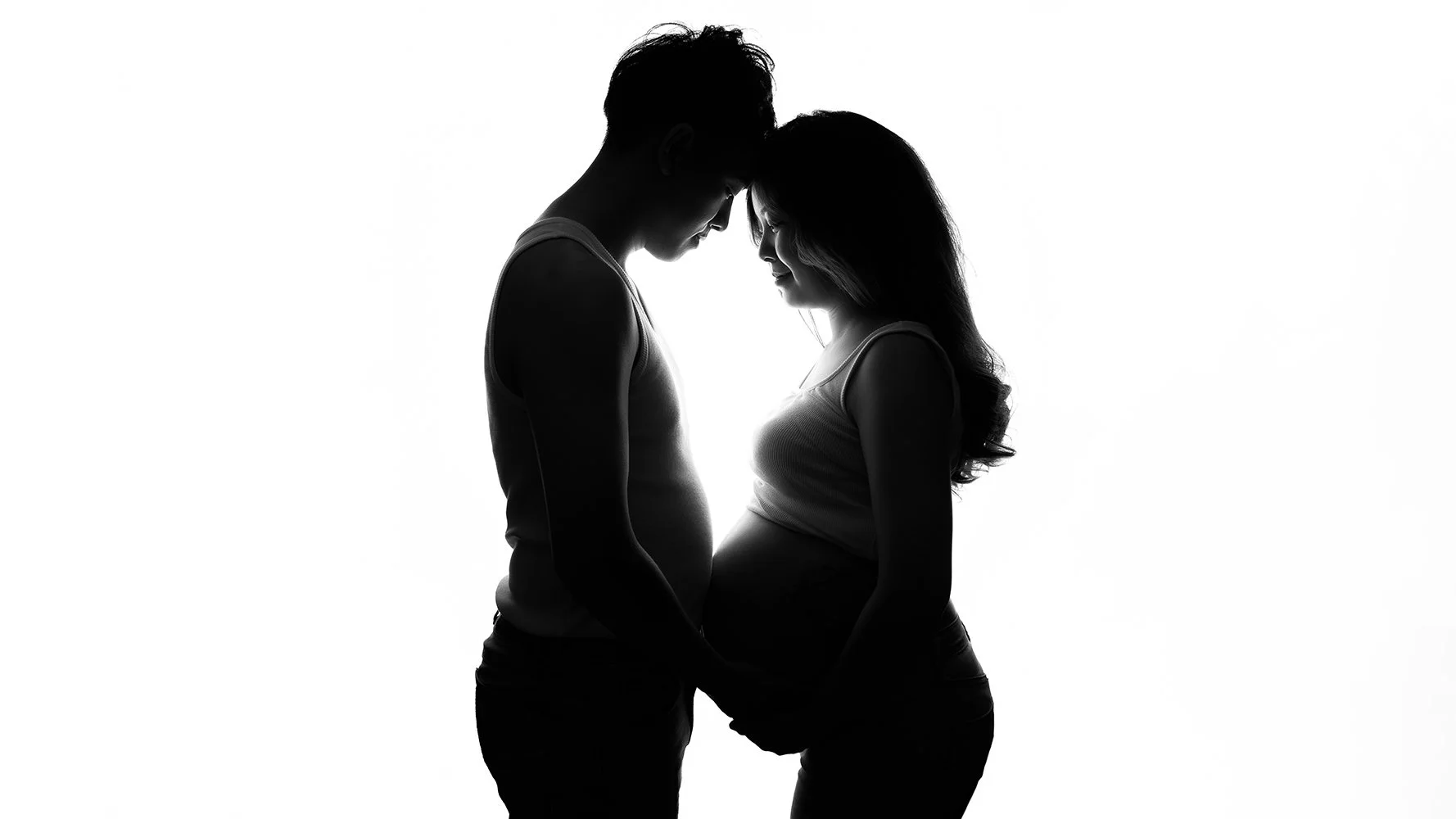 A low key silhouette portrait of a couple expecting their baby, standing next to each other looking at the belly