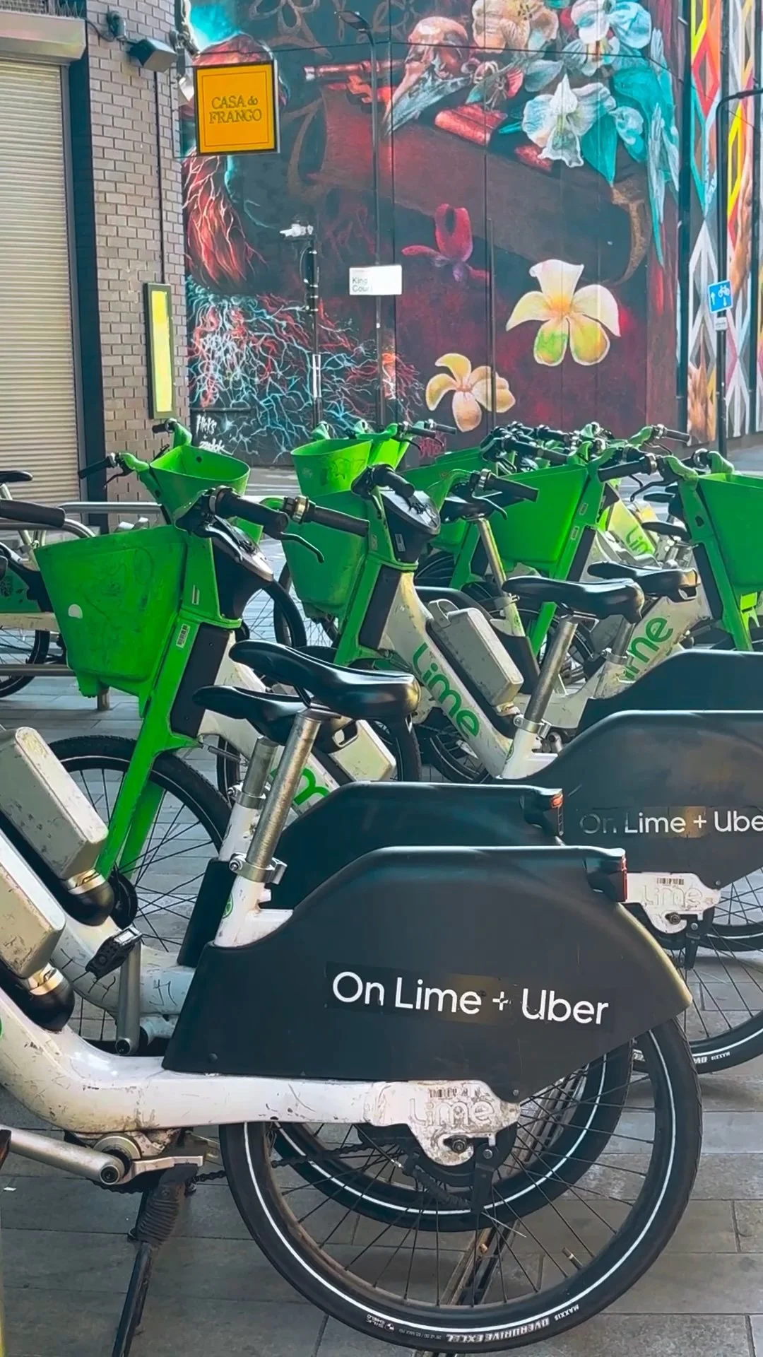 A line of green and black Lime electric bikes parked in front of a colorful graffiti mural on a building. The mural features various flowers and abstract designs.
