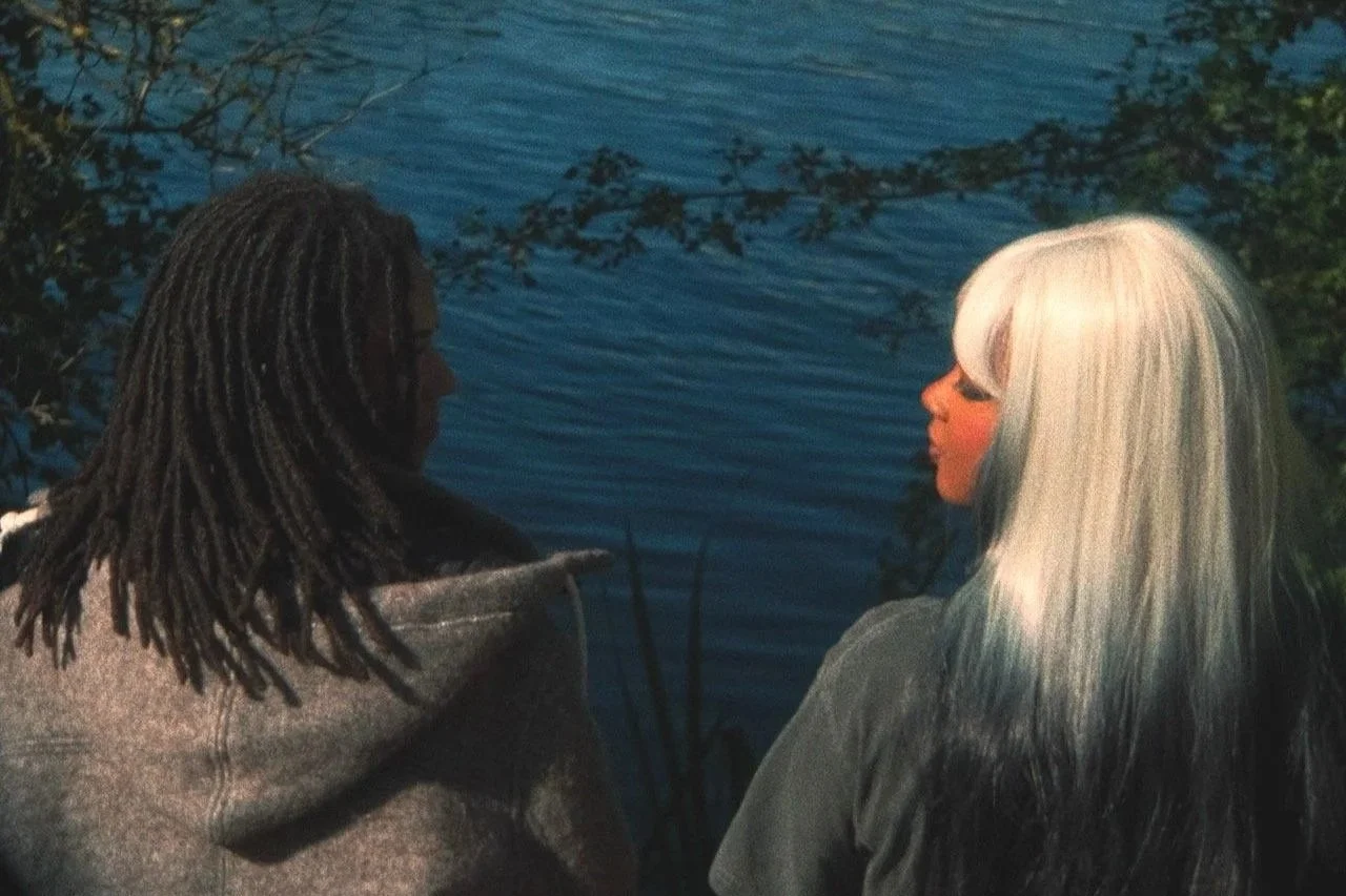 Two women with contrasting hair styles and colors standing near a body of water, surrounded by greenery, facing each other.