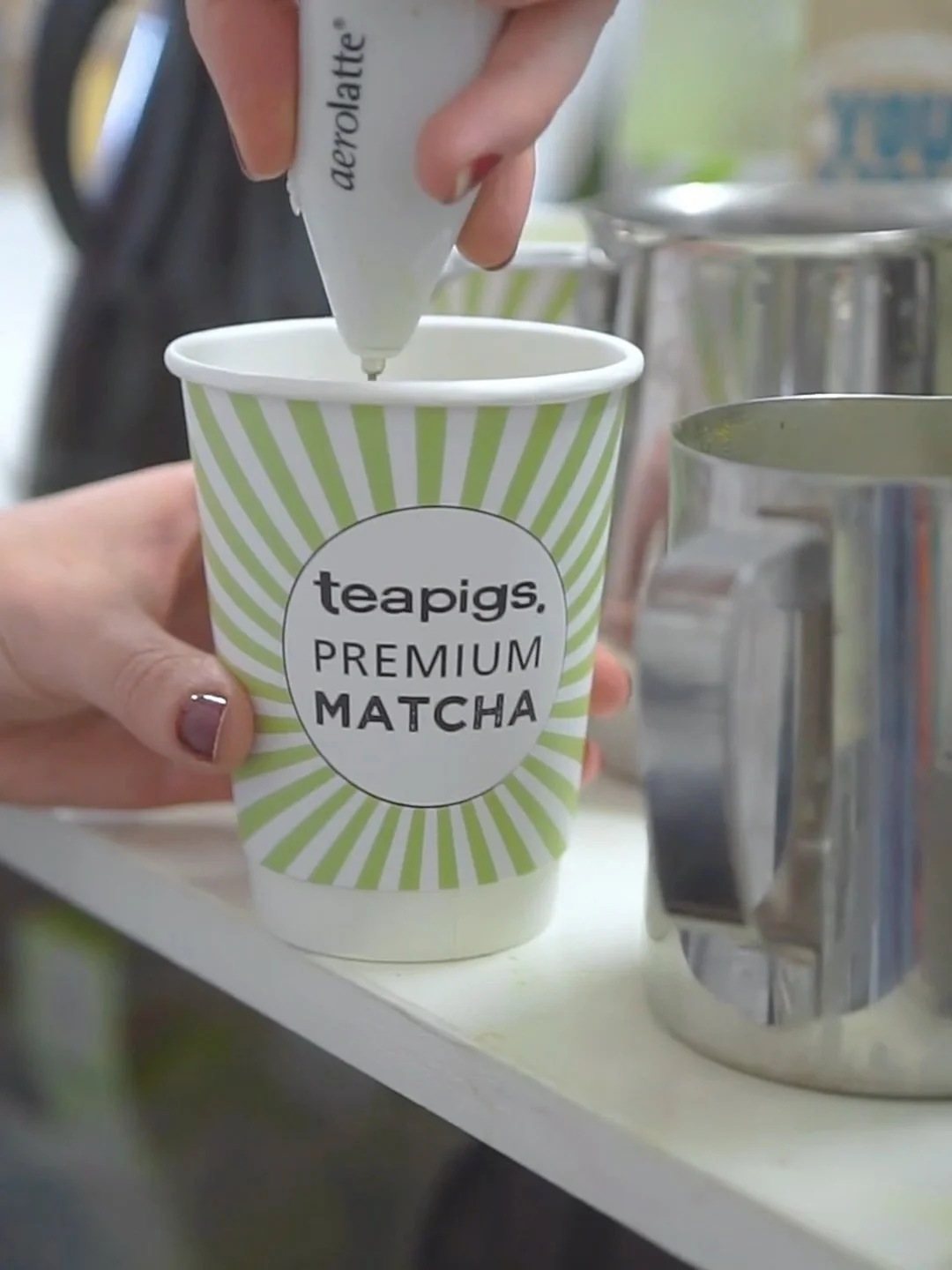 Person piping matcha powder from a white bottle with black text into a paper cup labeled 'teapigs premium matcha' with green and white radial stripes.