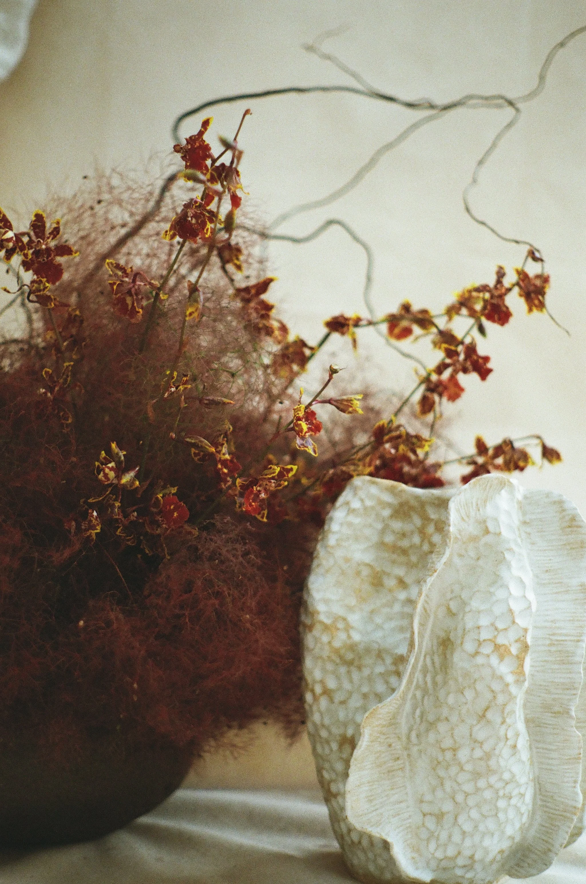 A close up image of a white ceramic vase with an organic shape and attached flat waves coming off of the surface from top to bottom with allover texture. A floral arrangement with purple sage bush and yellow and purple spotted orchids is in the back.