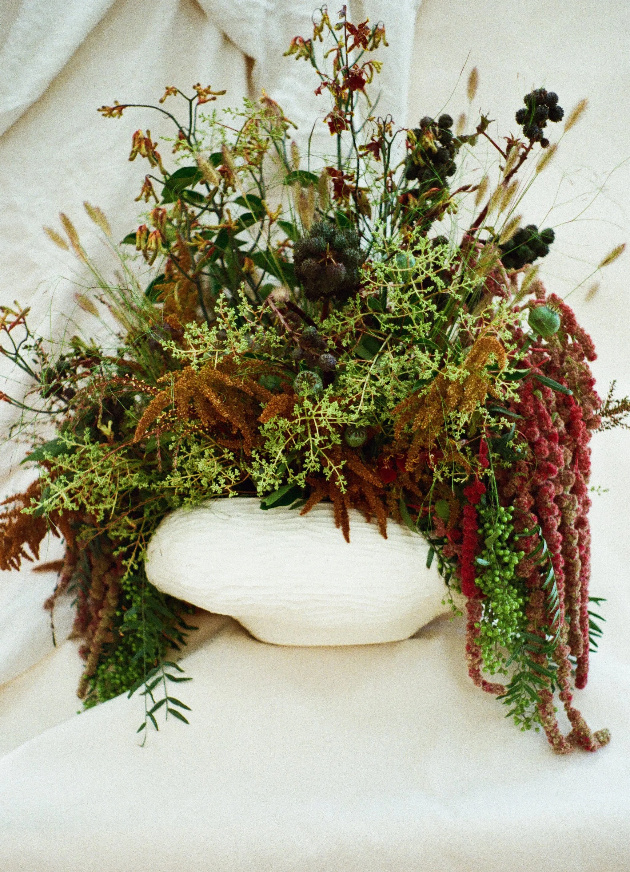 A large floral arrangement made up of green, orange, and dark pink flowers and leaves sitting inside of a large white handmade ceramic bowl with a carved wave like surface texture.