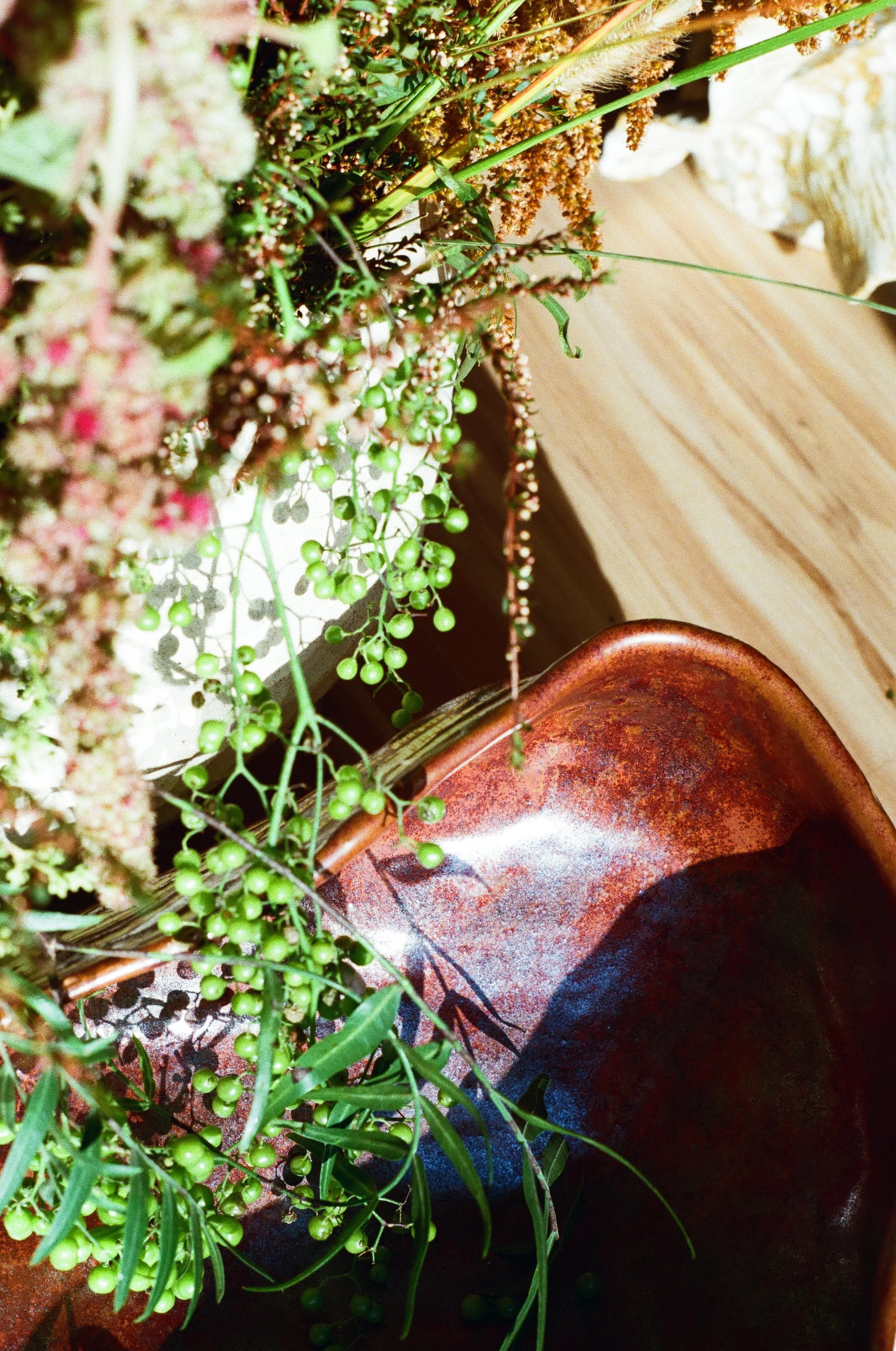 A close up image of a handmade ceramic bowl glazed with different shades of red with some green leaves spilling into it from a floral arrangement that is off camera.
