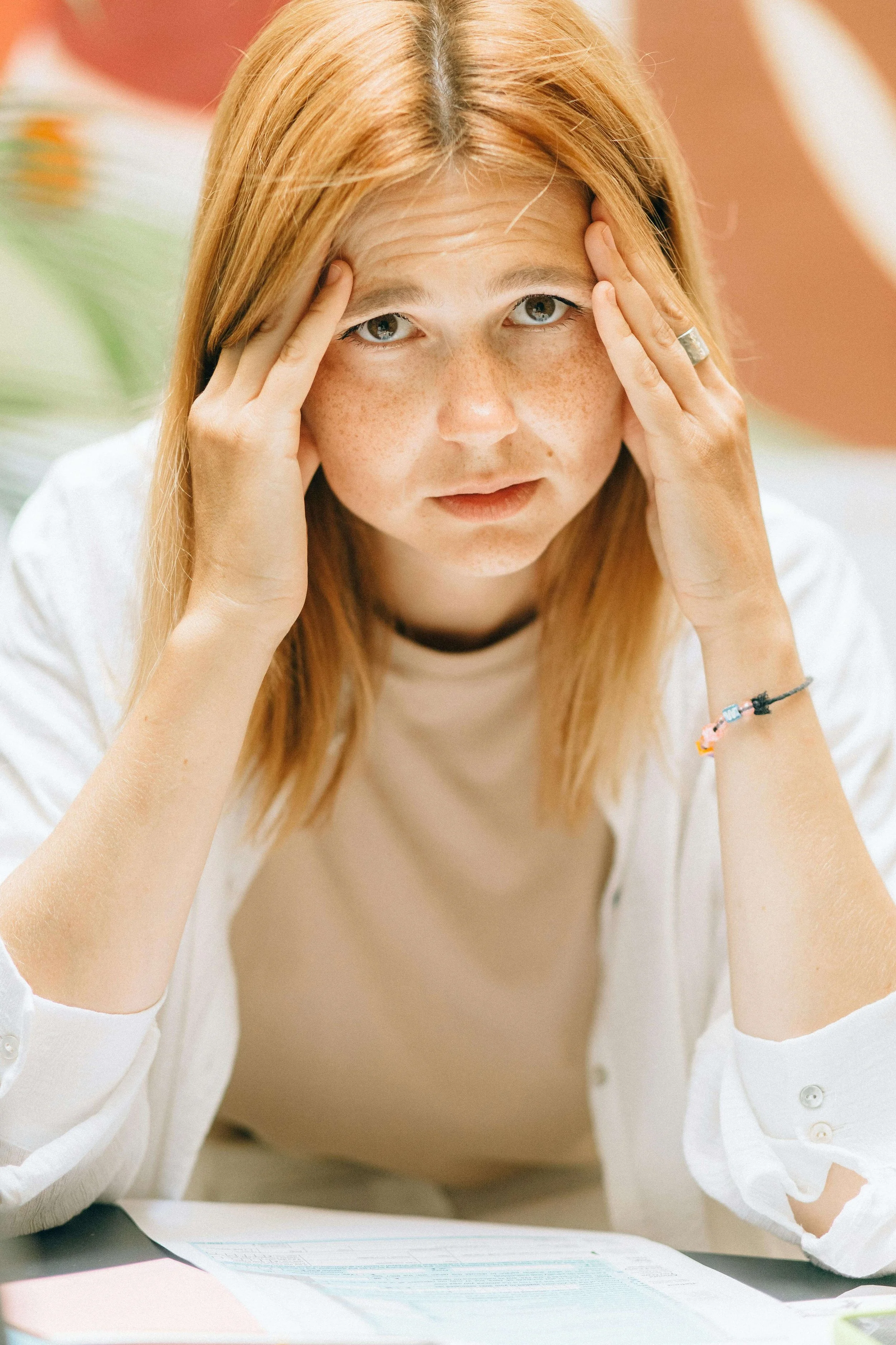 A young red haired woman with freckles holding both hands to her temples with a stressed expression. When anxious thoughts feel like they are taking over, anxiety therapy in Asheville, NC can help you examine them and finally take back control.