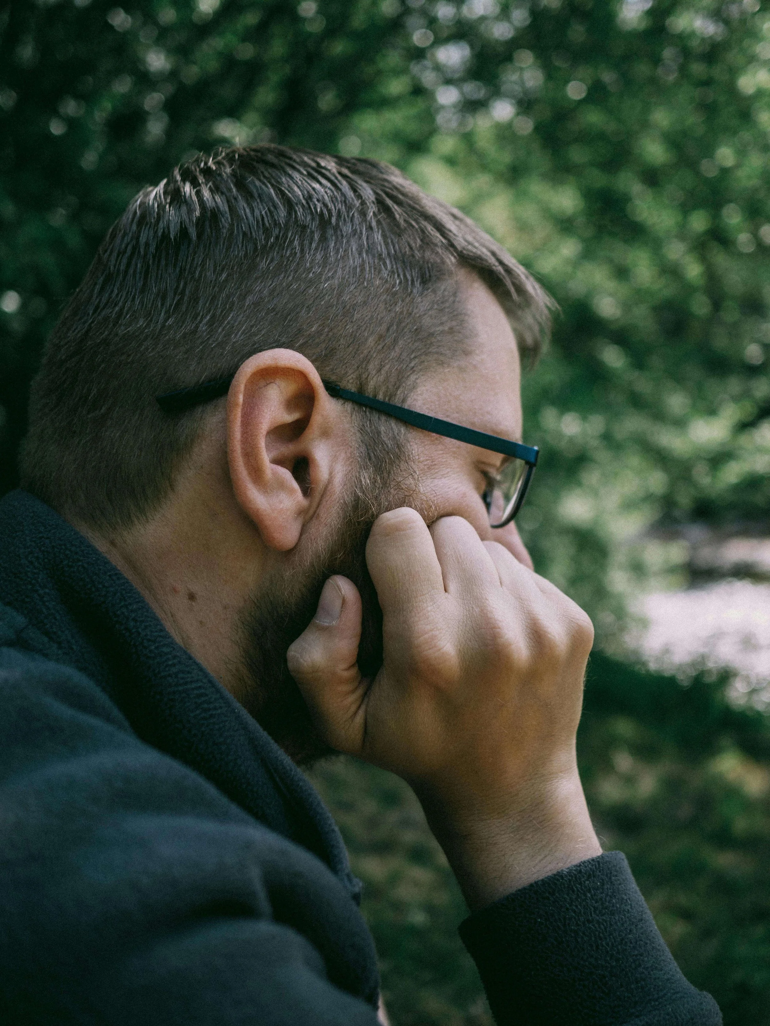 A bearded man wearing glasses and a dark hoodie standing outdoors with his hand pressed against his face. Worst case thinking doesn't have to run your life and anxiety therapy in Asheville, NC gives you the real tools to change that.
