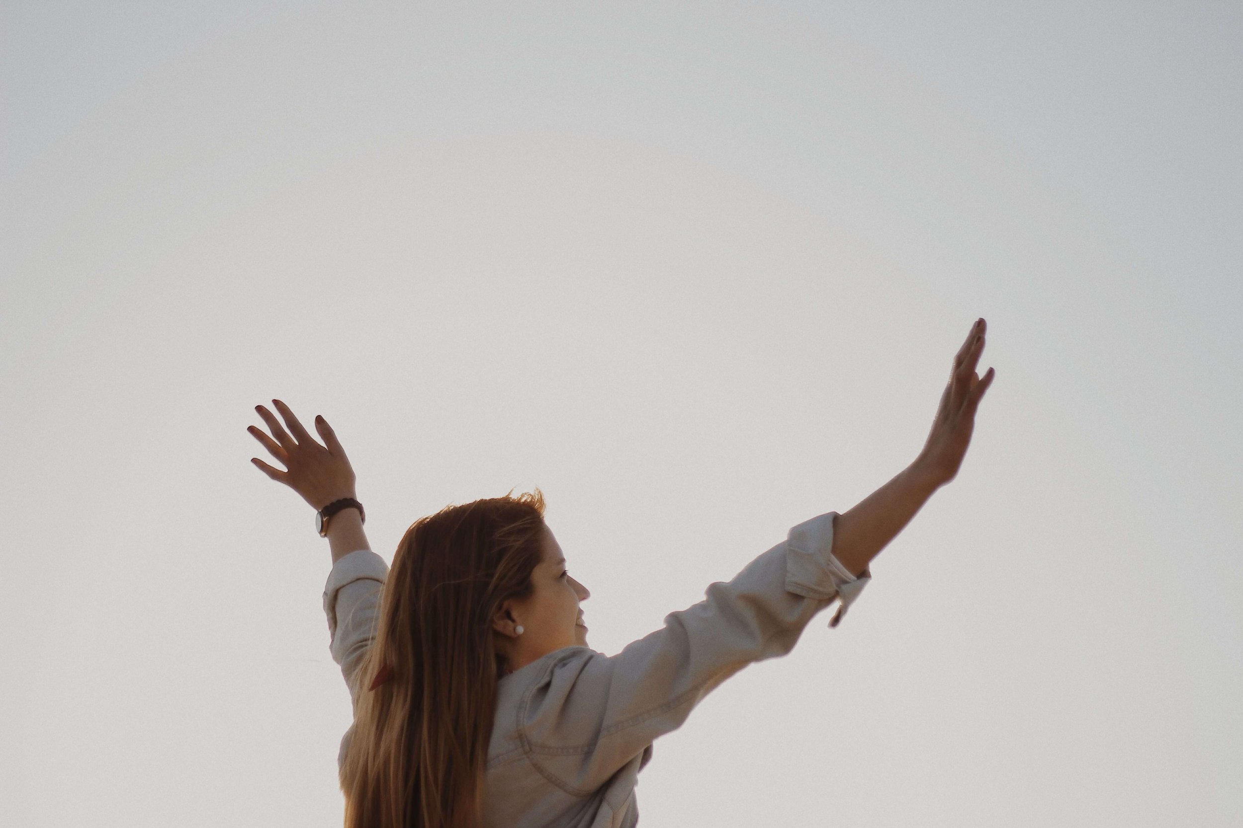 A woman with long hair wearing a jacket raising both arms wide open toward a gray sky. You don't have to wait for anxiety to disappear to start living and anxiety therapy in Asheville, NC can help you get there sooner than you think.