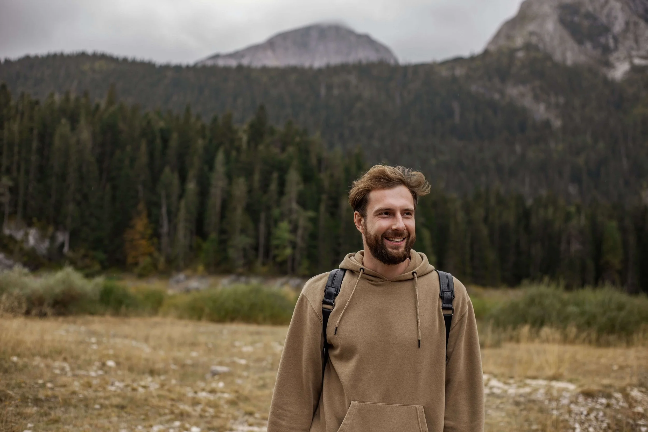 A smiling bearded man with a backpack standing outdoors in a grassy field with a dense forest behind him. Life beyond panic attacks is possible and anxiety therapy in Asheville, NC gives you the real tools to get there.
